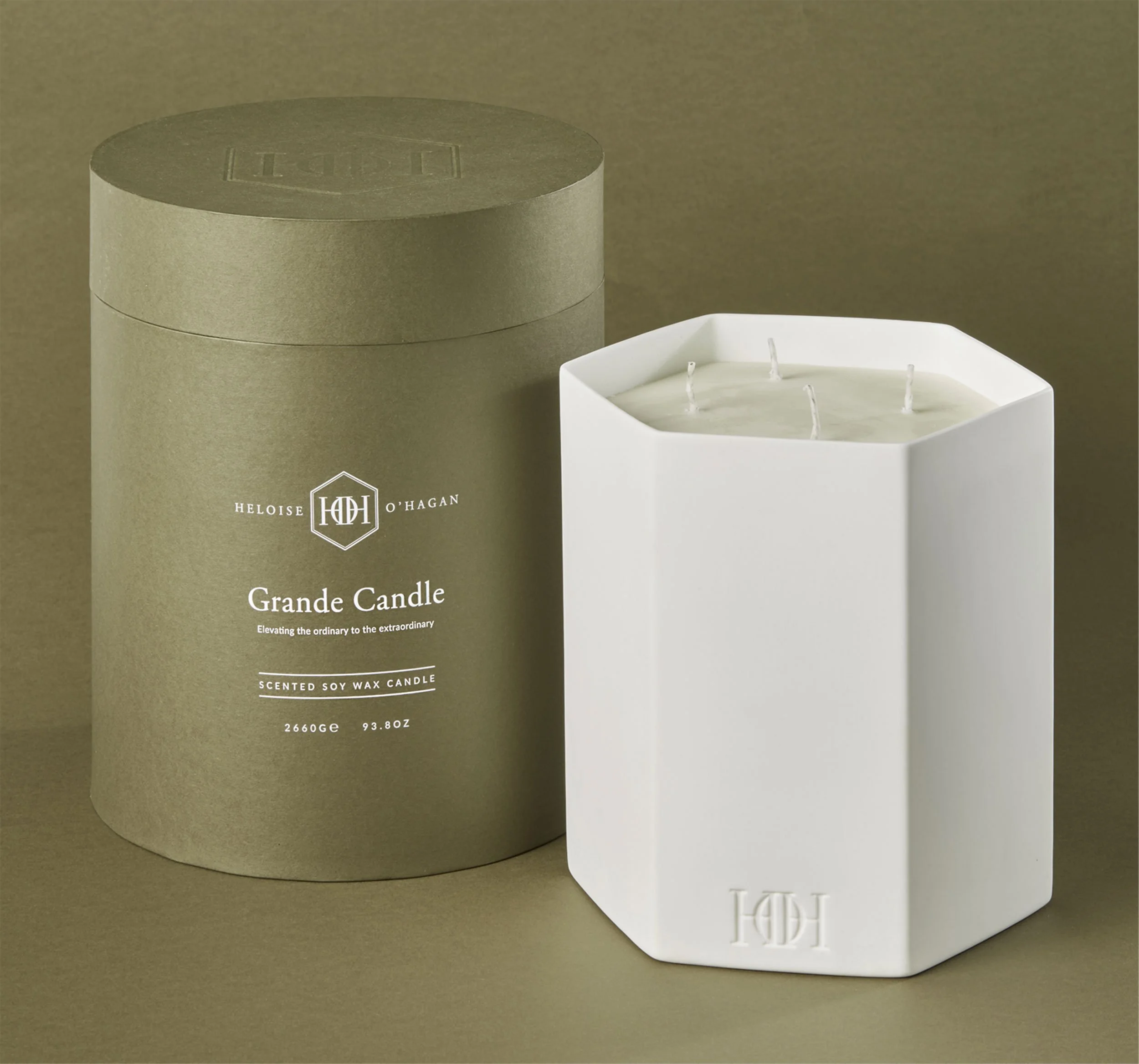 A white scented soy wax candle for Heloise O'Hagan, in a rectangular geometric holder with an embossed logo, beside a cylindrical box with the same logo, both on a beige background.