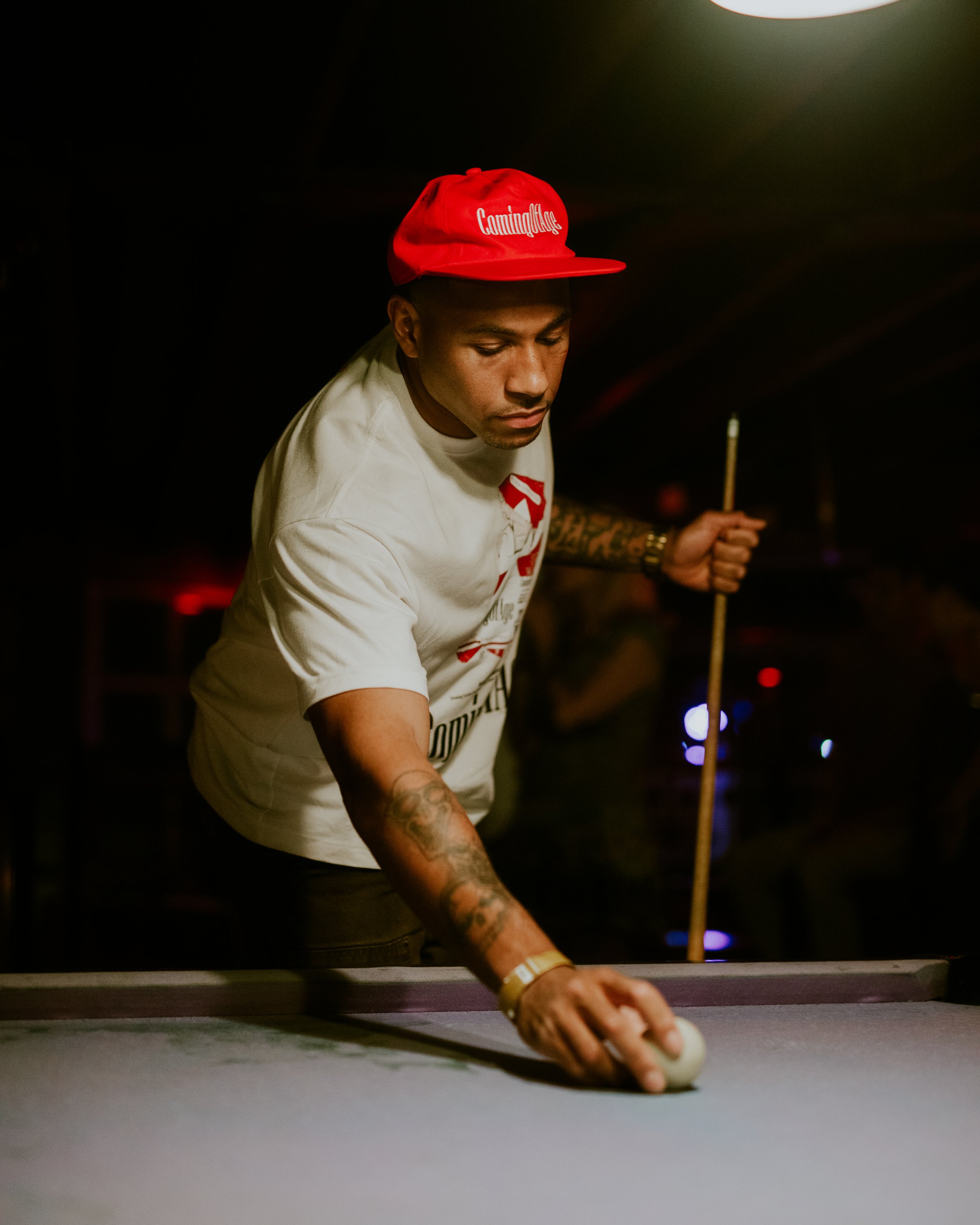 Young man in a white shirt and red cap playing pool in a dimly lit room.