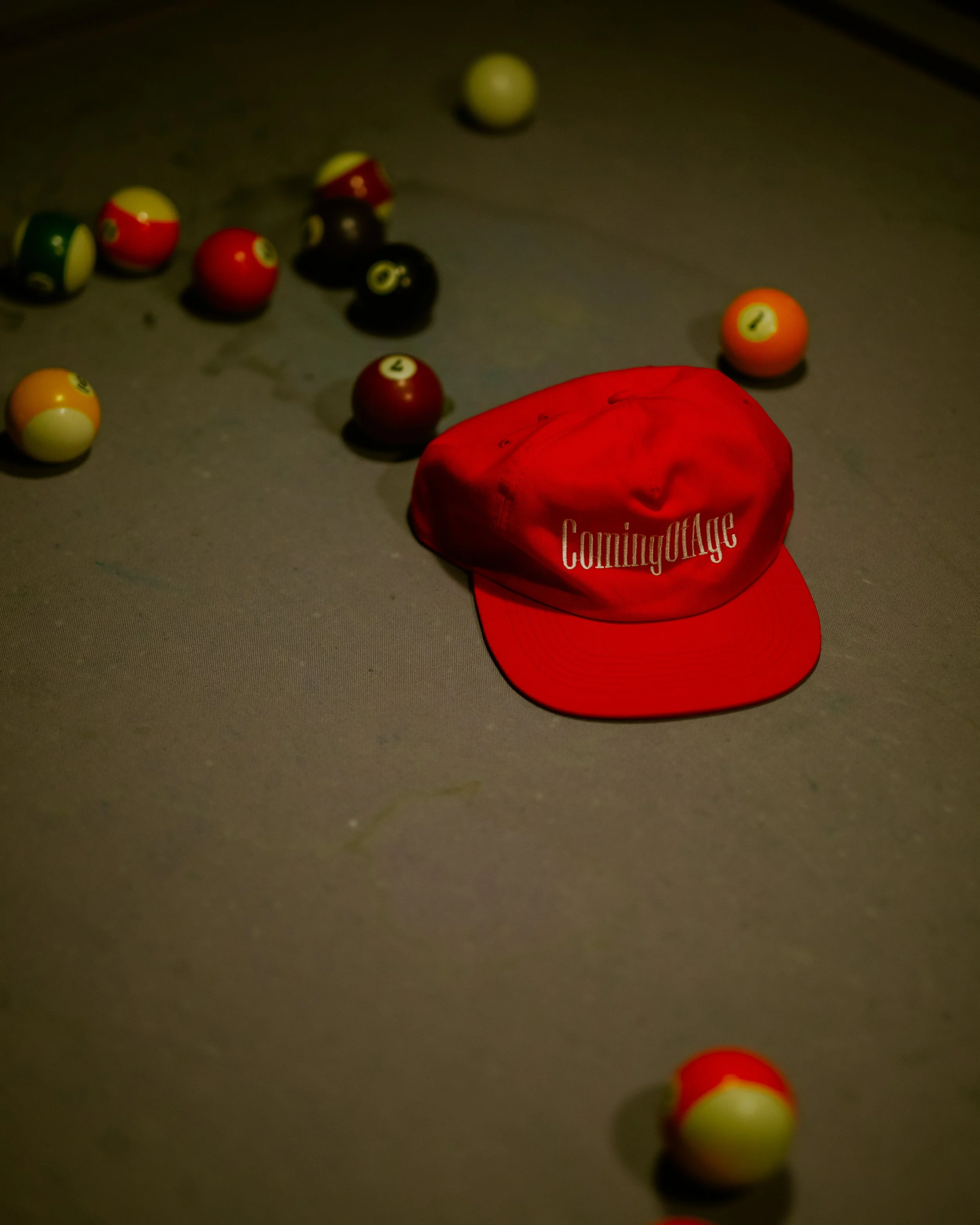A close-up of a pool table with several billiard balls scattered and a red hat with the words 'ComingOfAge' on it in the foreground.