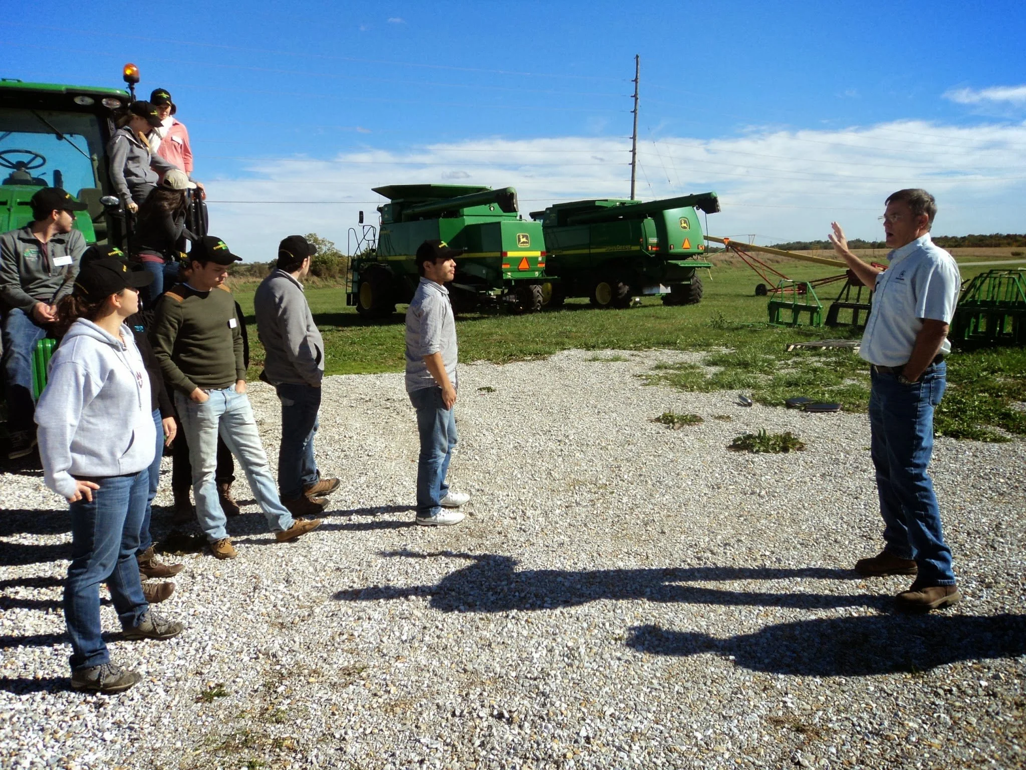 Fellows stand on a grave road in front of (and some standing on) John Deere farming equipment. They're listening to a man speak.