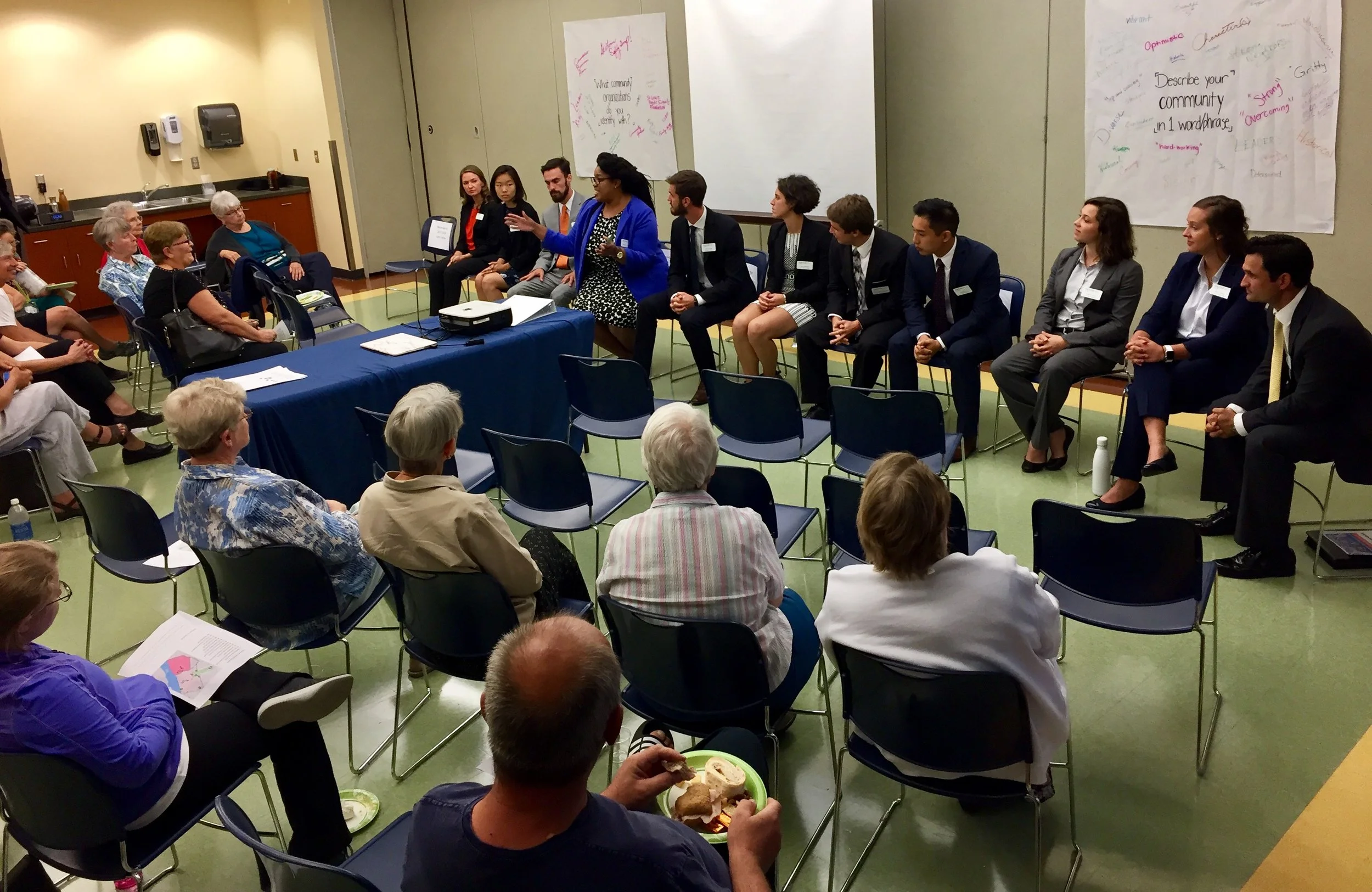 A group of fellows sits in front of a full room. They're listening to a woman wearing blue, sitting in the middle of the row of Fellows..