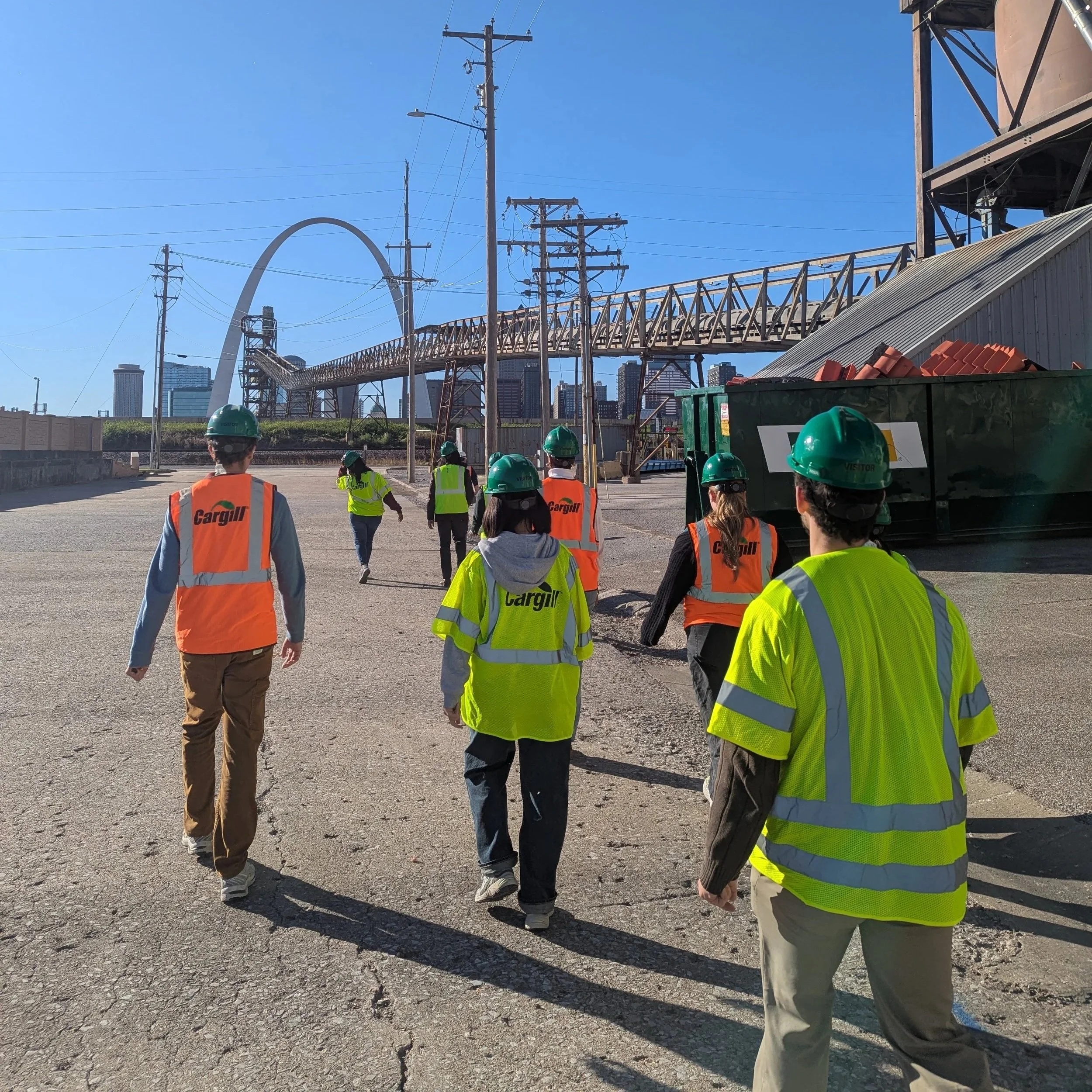 Fellows walk on a concrete walkway. There's a crane overhead and the St. Louis Arch is in the background.