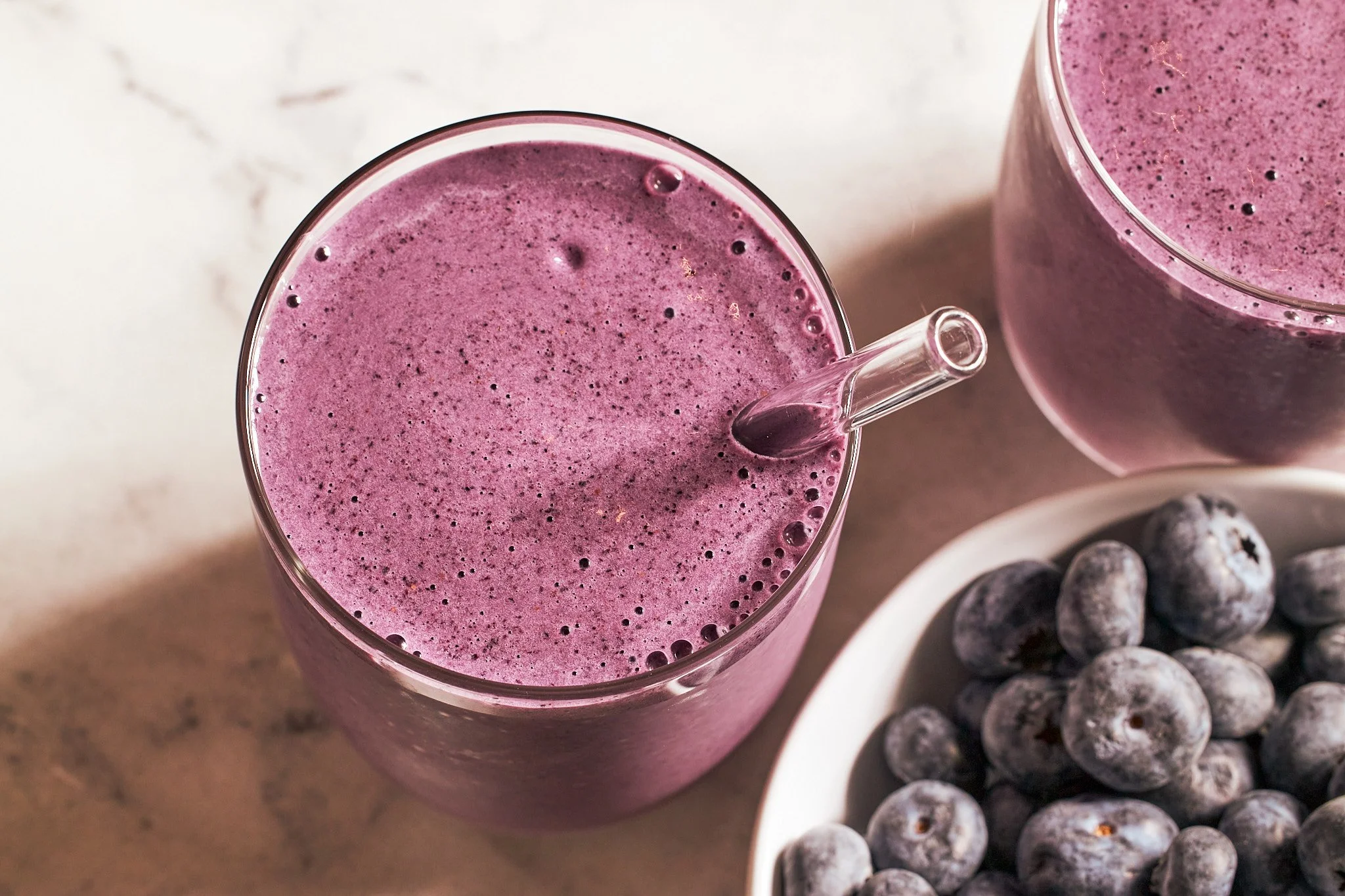 Blueberry smoothies in glass with fresh blueberries in a bowl on a neutral surface