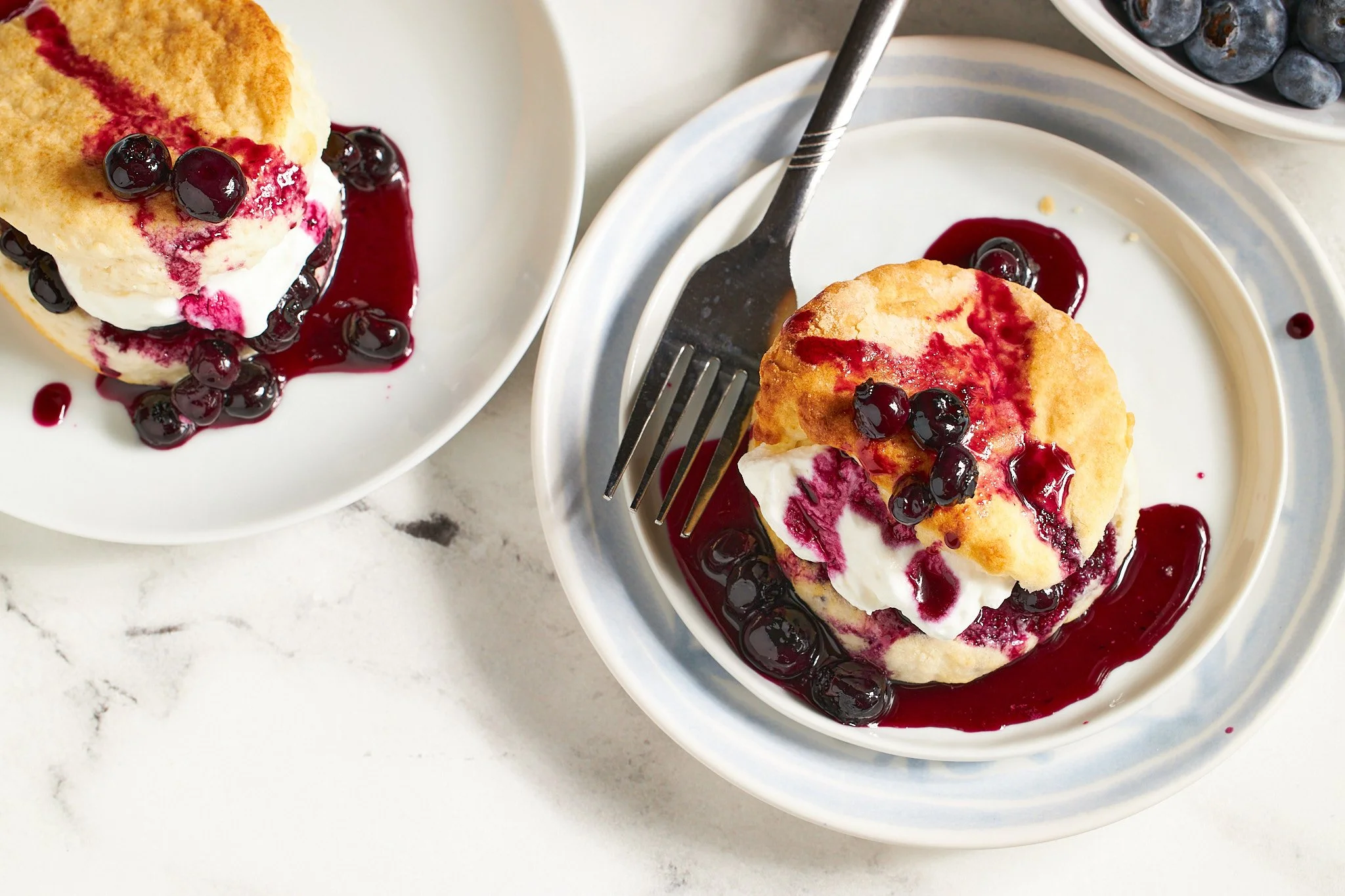 blueberry short cake with biscuit topped with blueberries in syrup and whipped cream with a fork on a blue plate with a white background