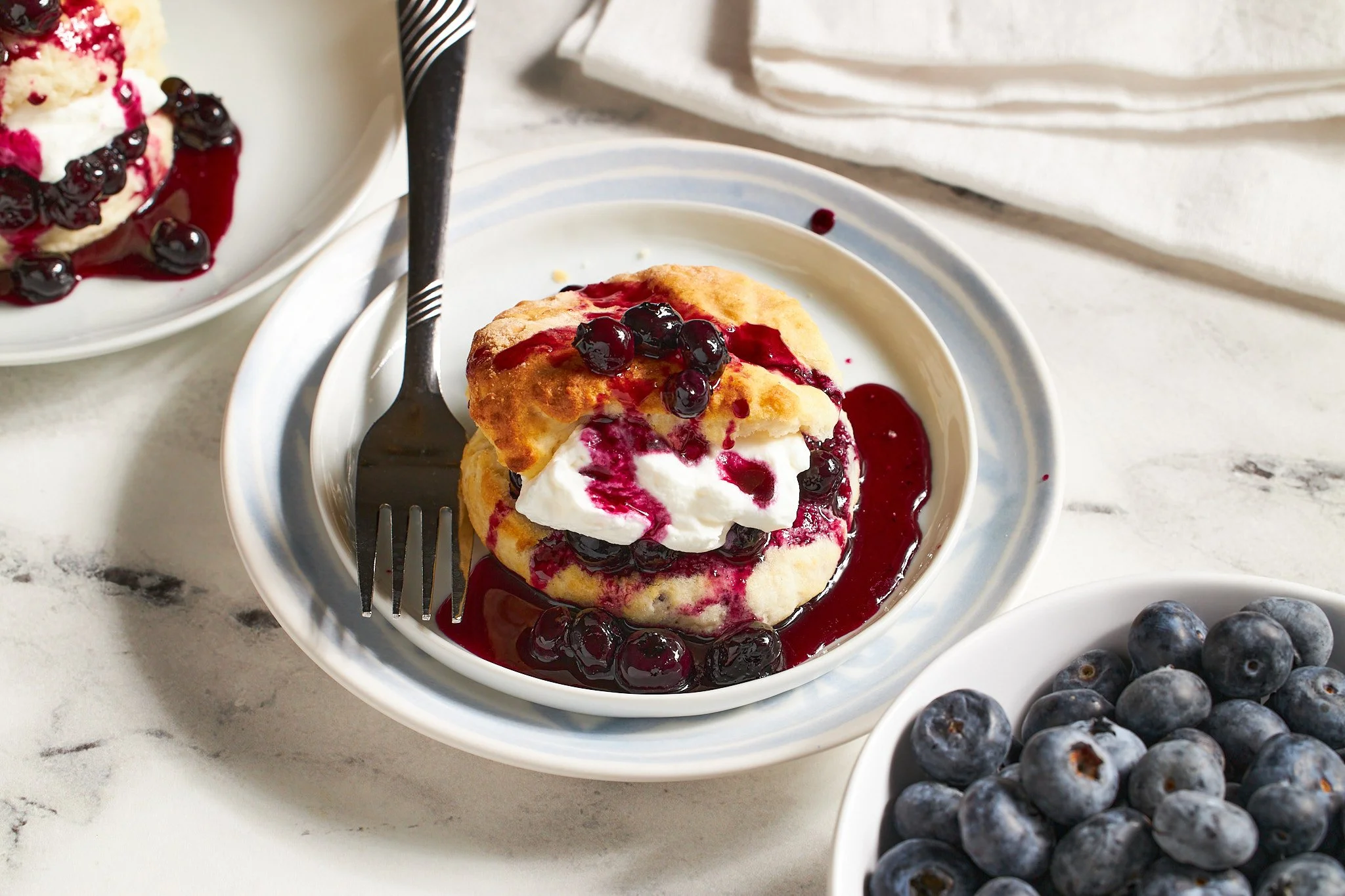 blueberry shortcake with biscuit topped with blueberries in syrup and whipped cream with a fork on a blue plate with a white background