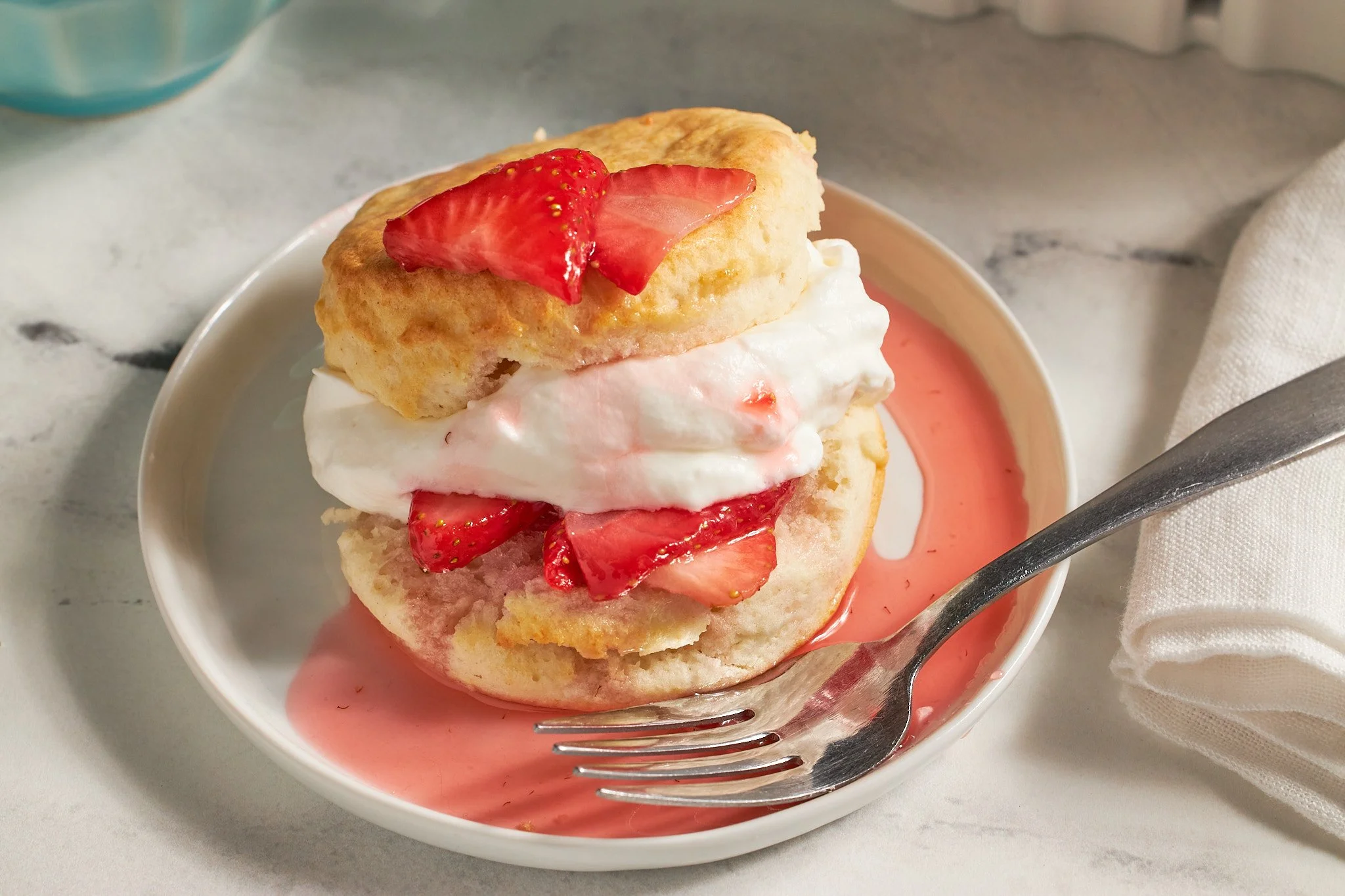 strawberry shortcake with biscuits fresh strawberries and whipped cream on a white plate with a fork on neutral background and aqua dishes