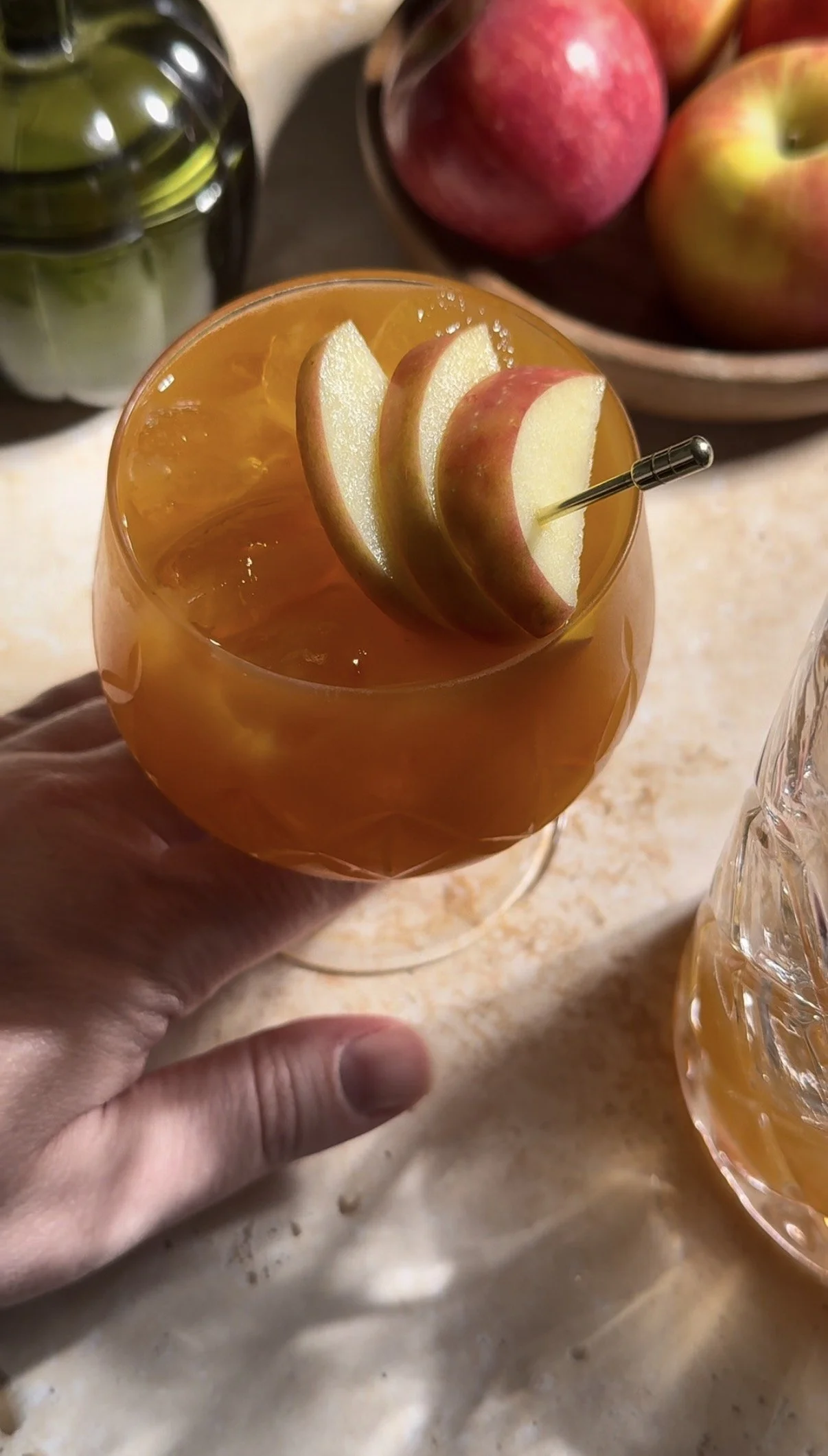 Photo of pumpkin butter apple mocktail in an etched glass with a garnish of apple slices on a golden toothpick with a bowl of apples in the background on a warm background with bright, direct light.