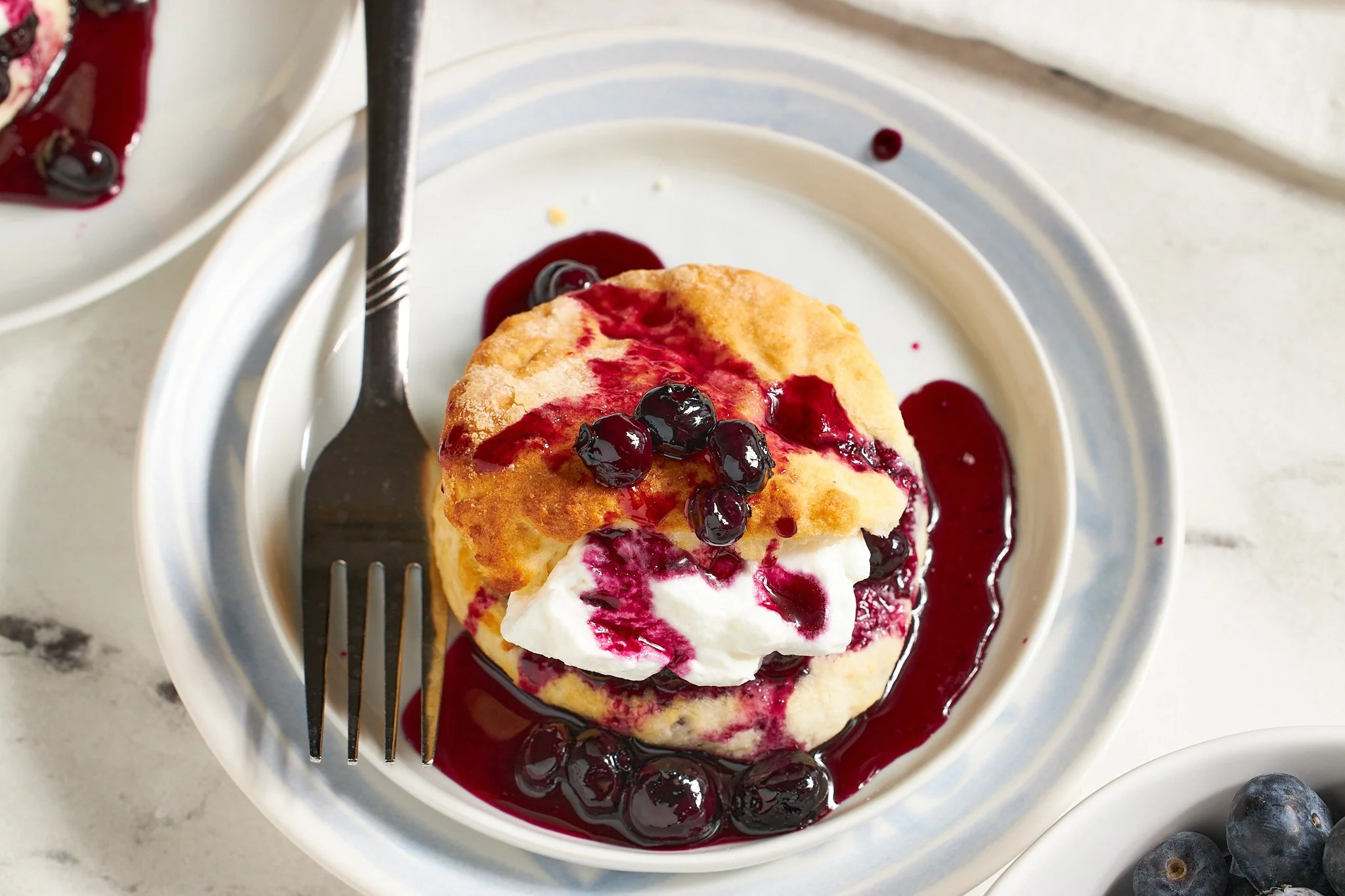 blueberry shortcake with biscuit topped with blueberries in syrup and whipped cream with a fork on a blue plate with a white background