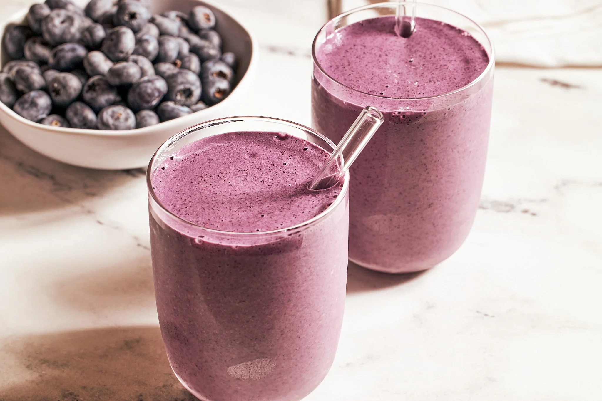 Blueberry smoothies in glass with fresh blueberries in a bowl on a neutral surface