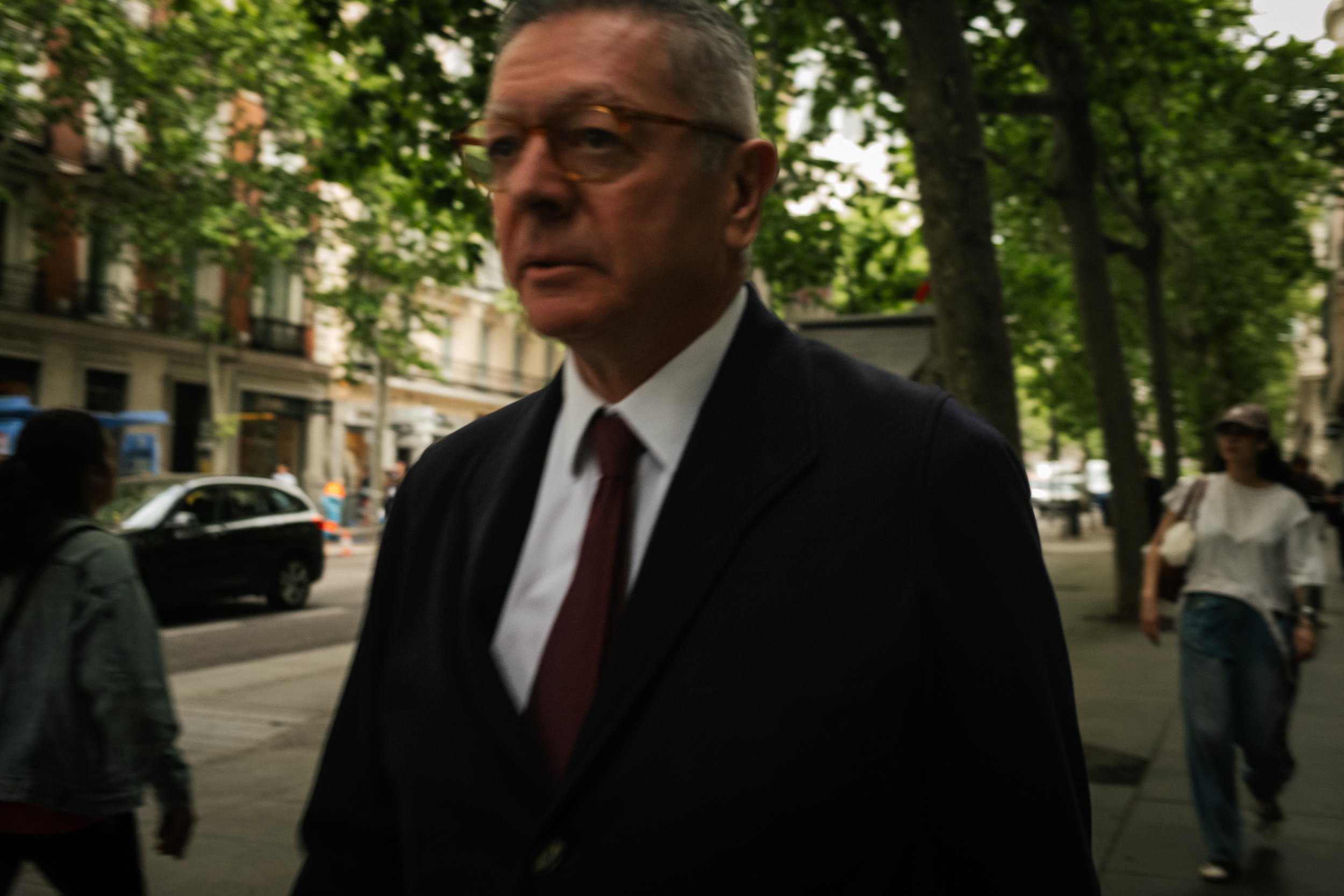 A man wearing glasses, a white shirt, a dark suit, and a red tie walking on a city sidewalk with green trees and buildings in the background.