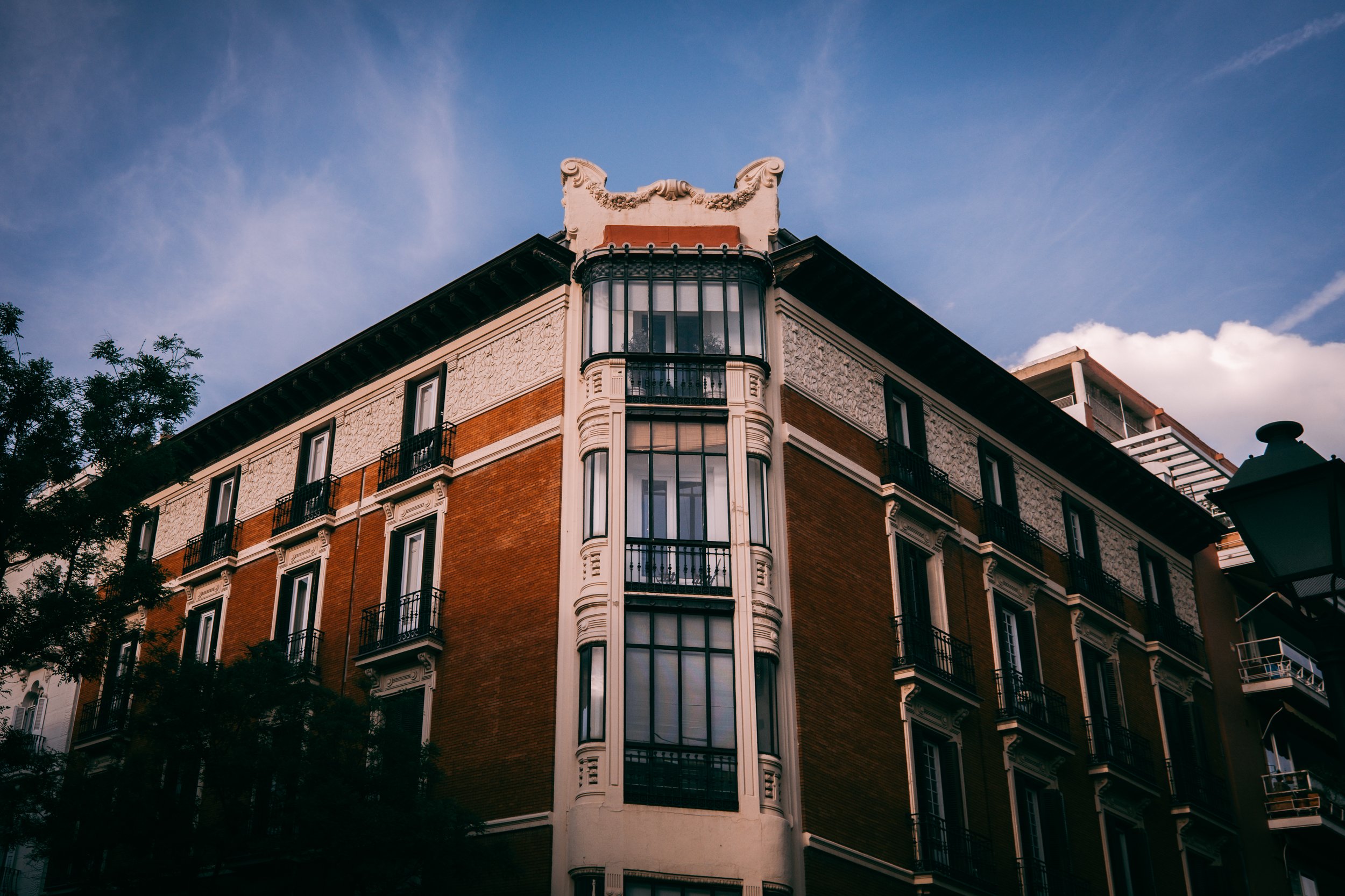 A multi-story building with ornate architectural details, including a curved window bay, decorative cornices, and balconies, against a blue sky with some clouds and trees at the bottom.