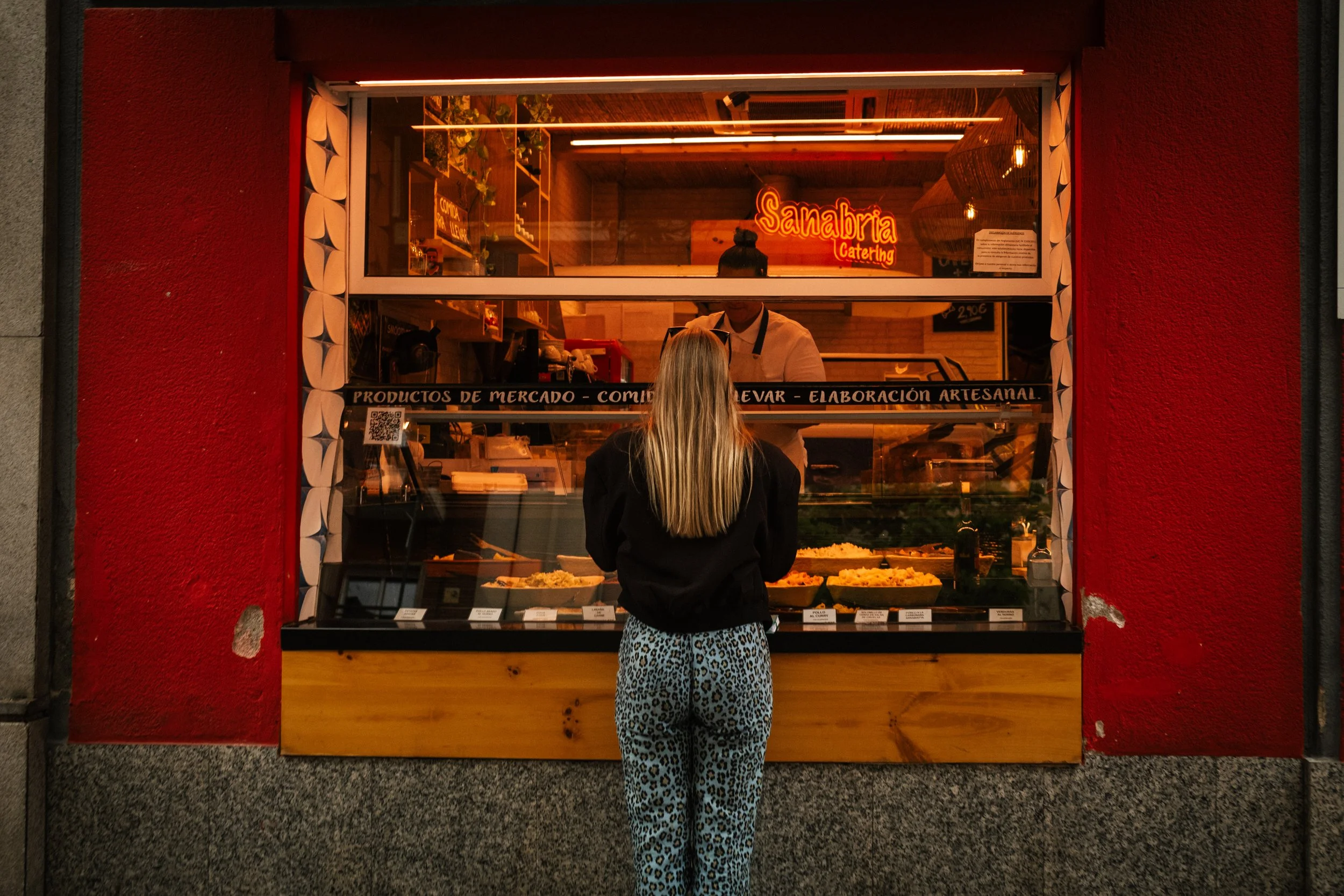 A woman with long blonde hair wearing leopard print pants and a black jacket is standing in front of a food stall called Sanabria Catering. The stall has a red exterior wall, a large window displaying various food items, and a neon sign with the name