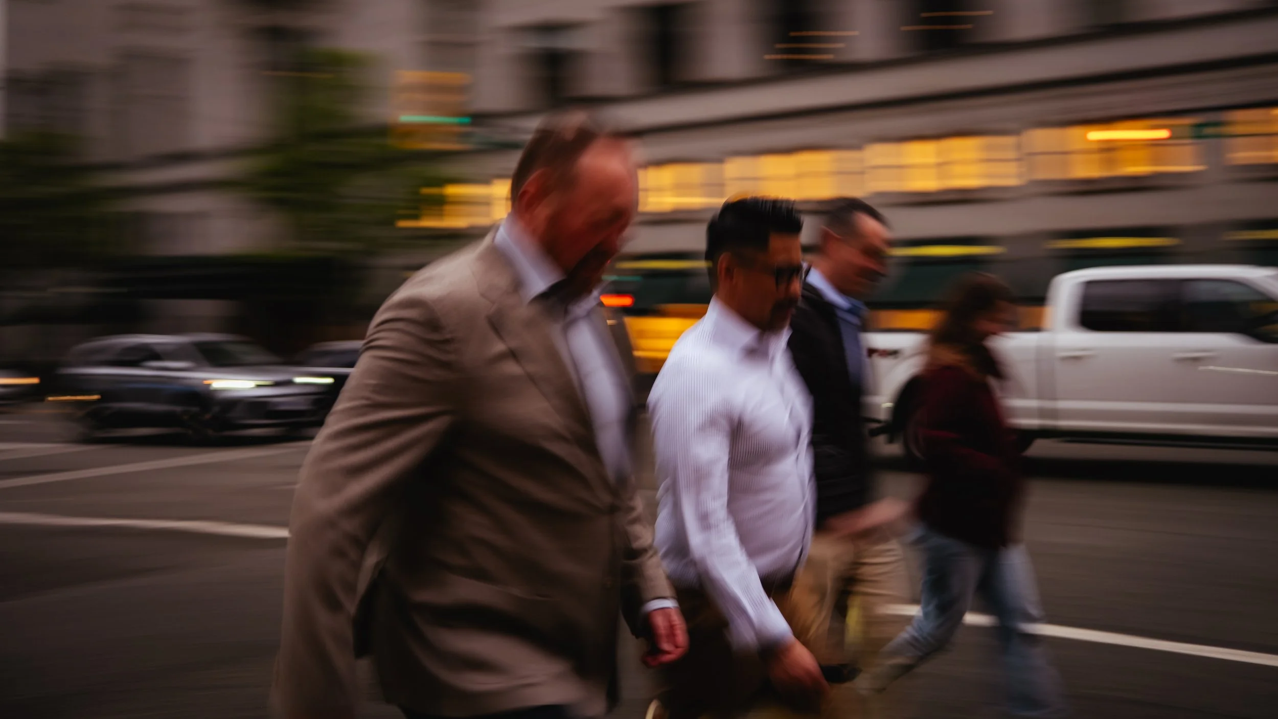Group of people walking on city street with blurred cars and buildings in background.