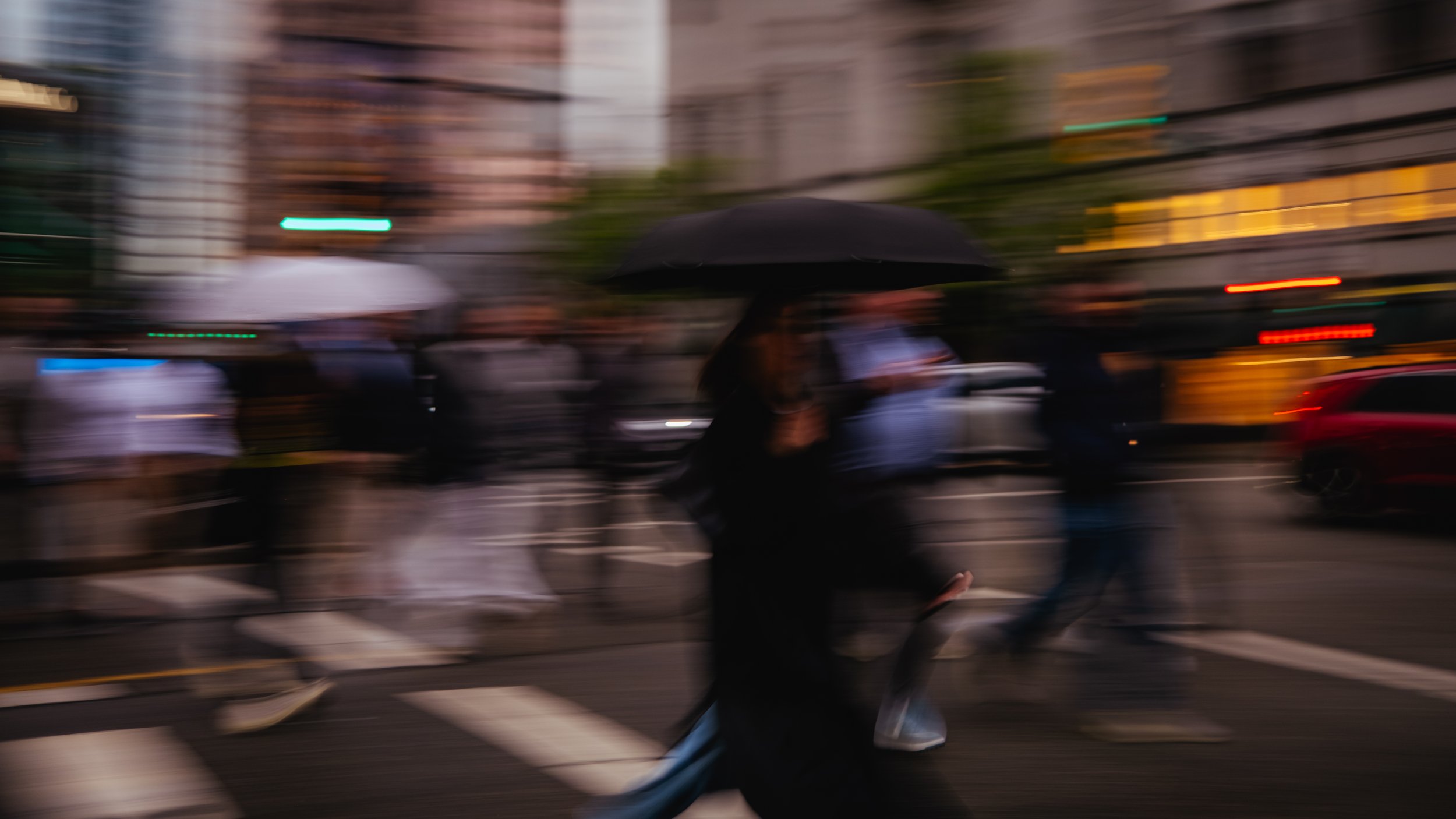 Blurry city street scene with pedestrians crossing in rain, holding umbrellas, cars, and buildings in the background.