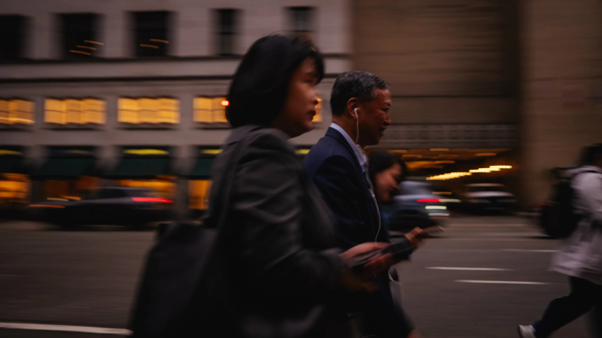 People walking on a city street at dusk with blurred background buildings and moving cars.