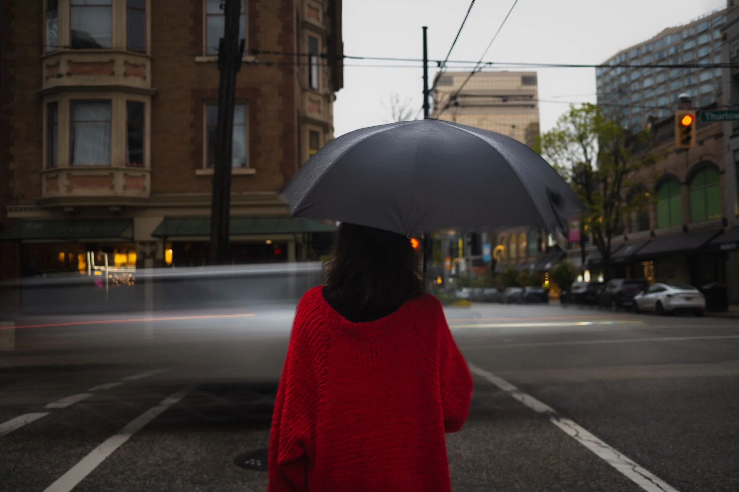 A person wearing a red sweater holding a black umbrella, standing on a city street during rainy weather, with buildings, trees, and traffic lights in the background.