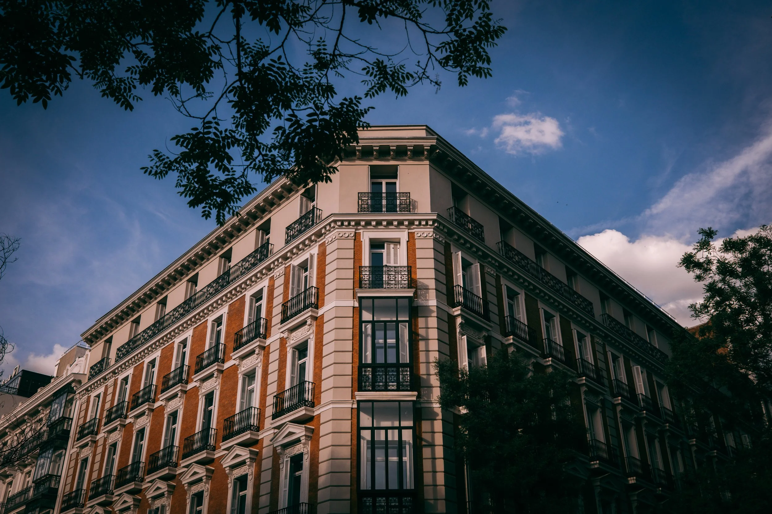 Photo of a multi-story residential building with white and orange brick exterior, black balconies, and large windows, under a partly cloudy blue sky, with tree branches in the foreground.