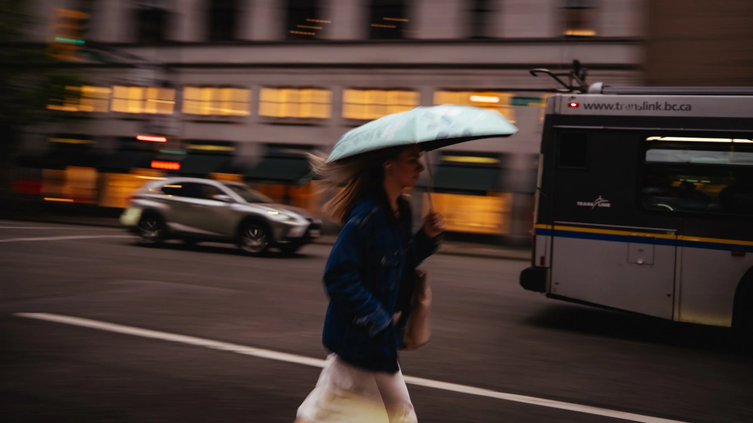 A woman walking on a city street at night, holding an umbrella, with cars and a bus passing by.