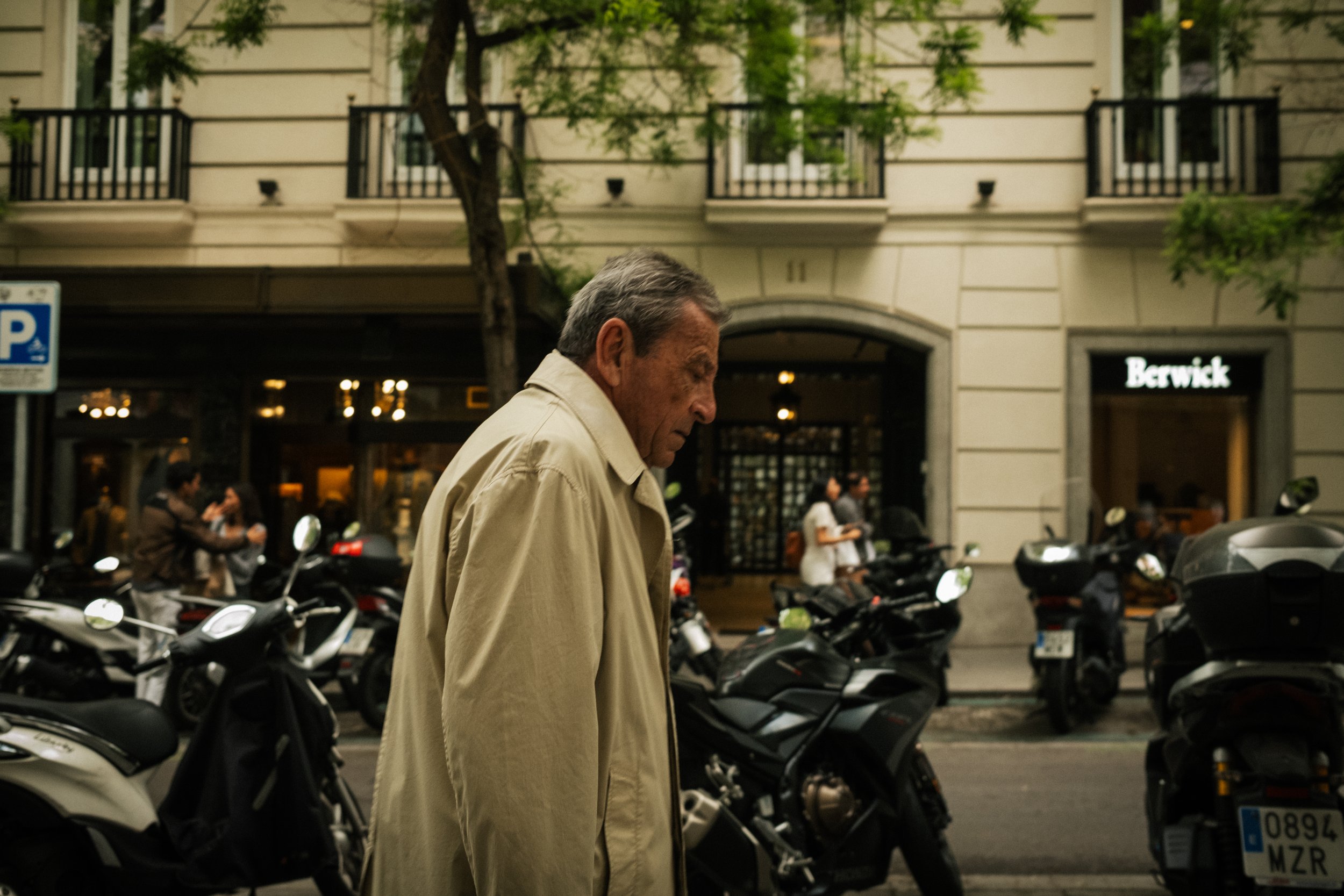 An elderly man walking past parked scooters on a city street with storefronts and a building with balconies in the background.