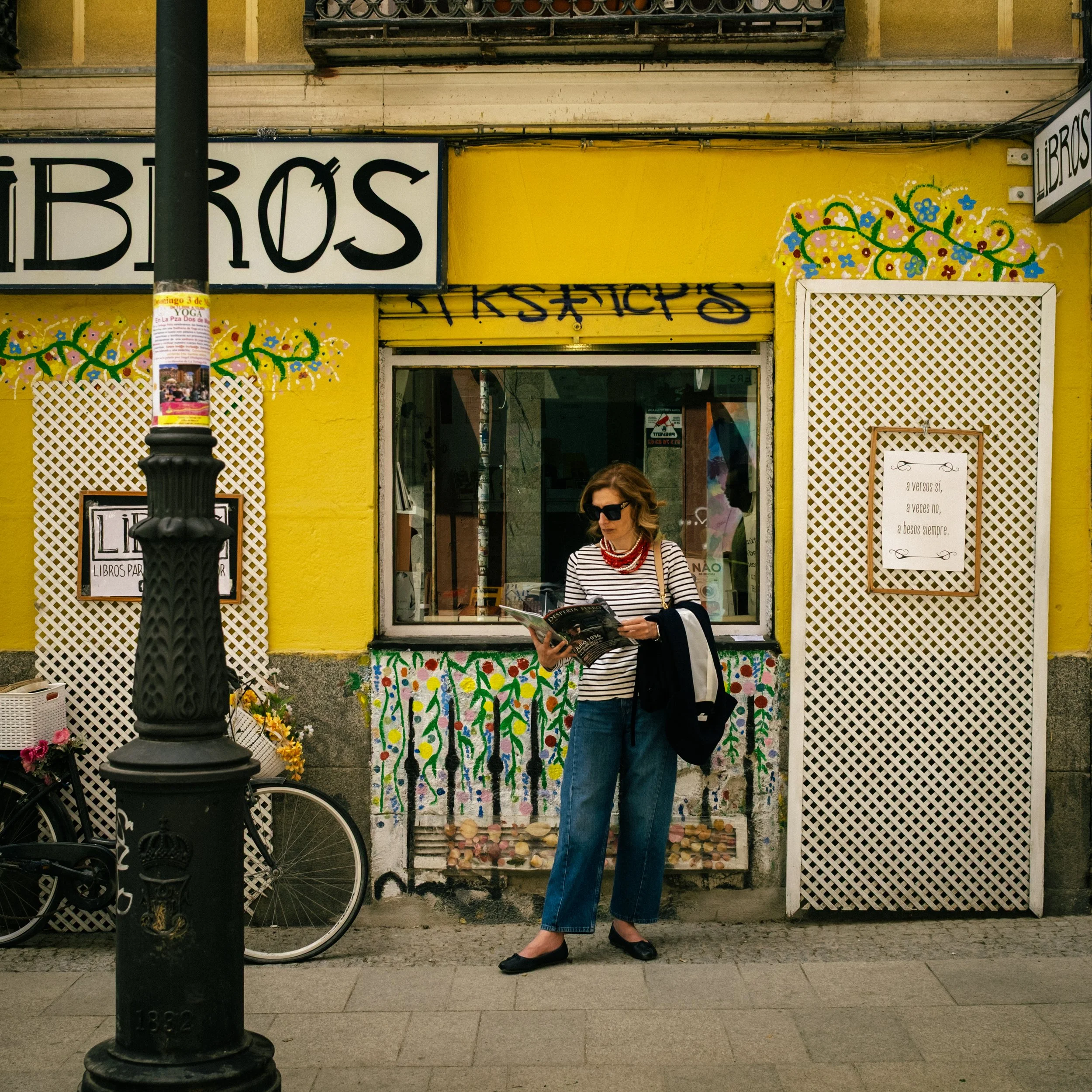 A woman standing in front of a yellow storefront wall with colorful floral artwork, reading a magazine, wearing sunglasses, a striped top, jeans, and black shoes, partially obscured by a black lamppost and a bicycle.