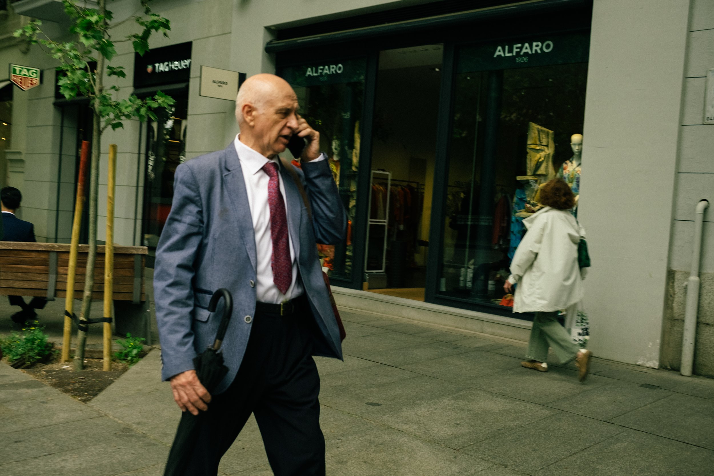 A man in a gray suit and pink tie walking on a city sidewalk, talking on his cellphone, with store windows and pedestrians in the background.