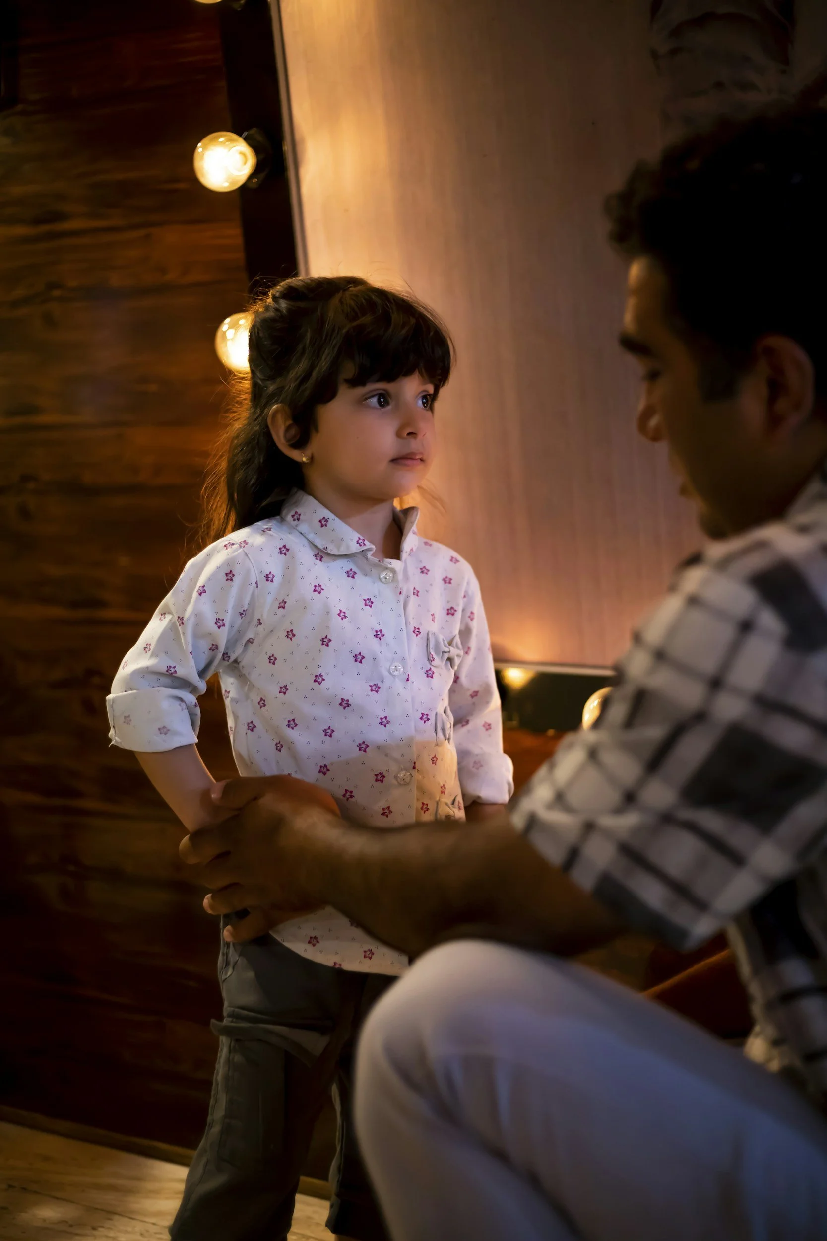 A young girl with dark hair, wearing a white shirt with small pink flowers, stands with her hand on her hip in a warmly lit room, holding hands with an adult man who is seated and wearing a striped shirt.