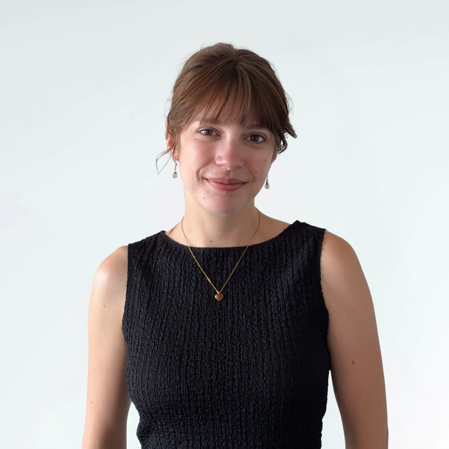 A woman with short brown hair and light skin, smiling at the camera, wearing a black sleeveless textured dress, green earrings, a gold necklace, and a heart-shaped pendant necklace, standing against a plain white background.
