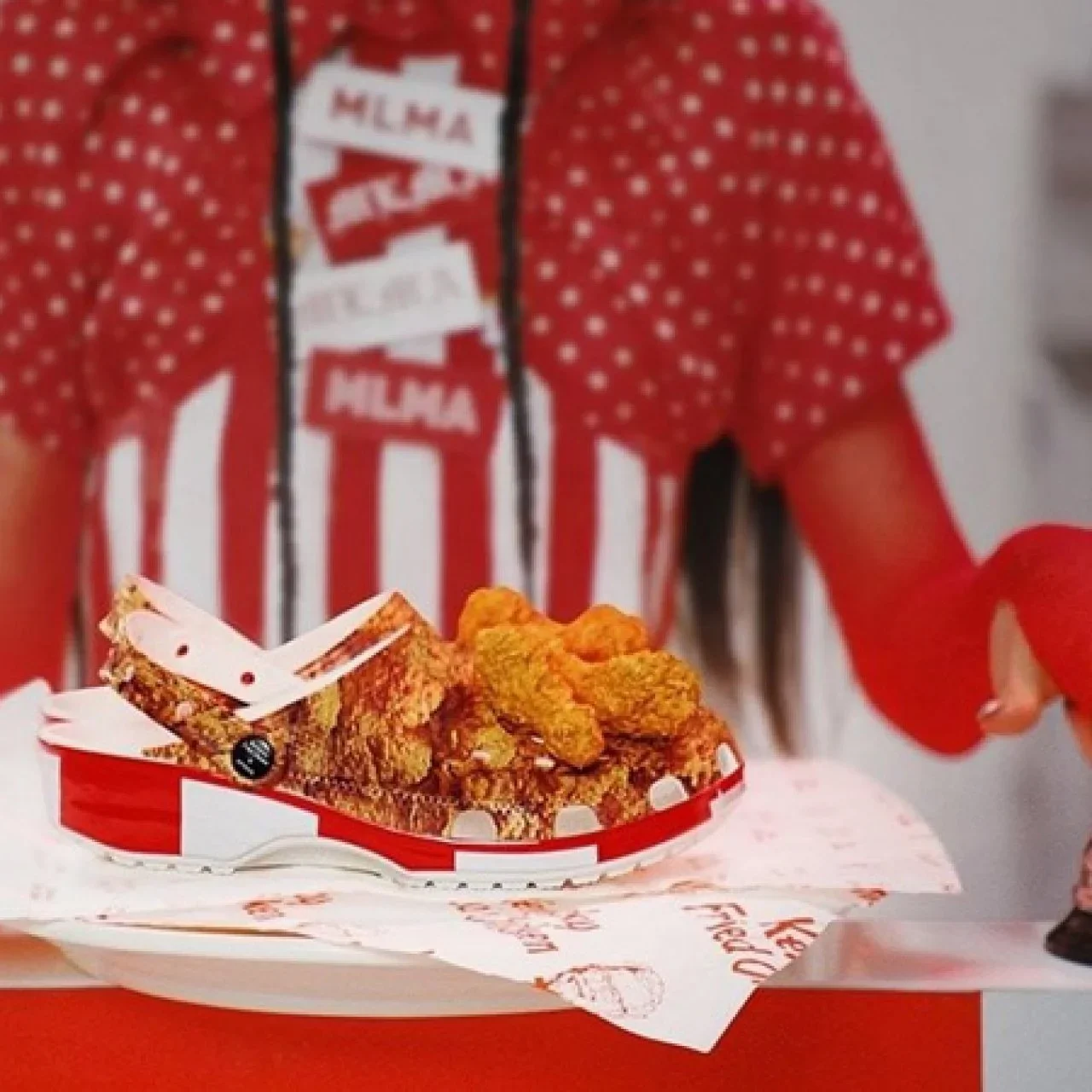 A hand holding a tray with fried chicken pieces on a red and white checkered paper, with a person in the background wearing a red and white striped shirt.