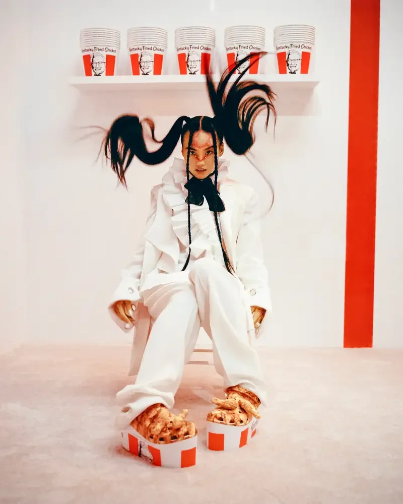 Person with long braided hair, dressed in white, sitting in a white room with a red stripe on the wall. They are wearing oversized fried chicken-themed shoes and sitting in front of a shelf filled with buckets of KFC fried chicken.