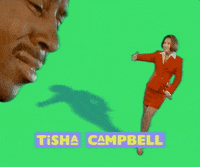 A promotional image for Tisha Campbell featuring her standing on a green background, pointing to the side with her right hand, wearing a red suit and short hair, with a shadow cast on the background.