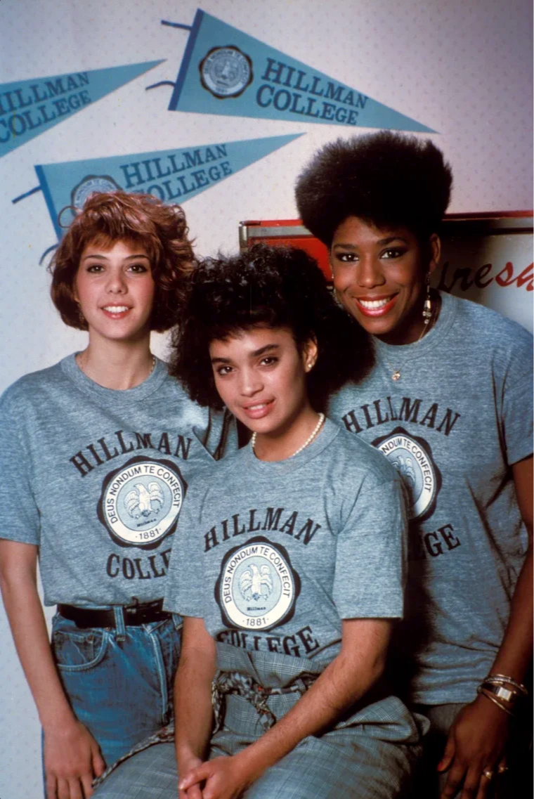 Three young women wearing gray Hillman College t-shirts, posing together in front of blue Hillman College pennants on a wall.