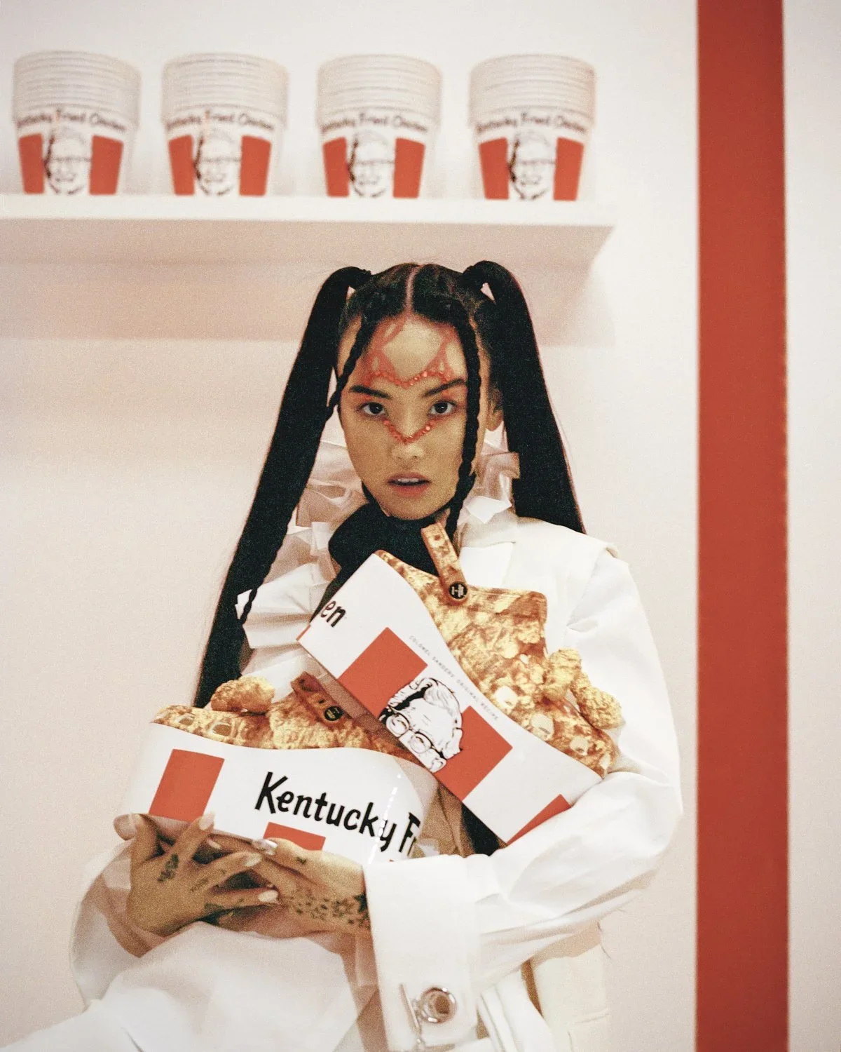 A woman with long black braided hair and facial makeup in a distinctive pattern, wearing a white jacket, holding a box and fried chicken on a tray from Kentucky Fried Chicken, standing in front of shelves with KFC cups, inside a KFC restaurant.