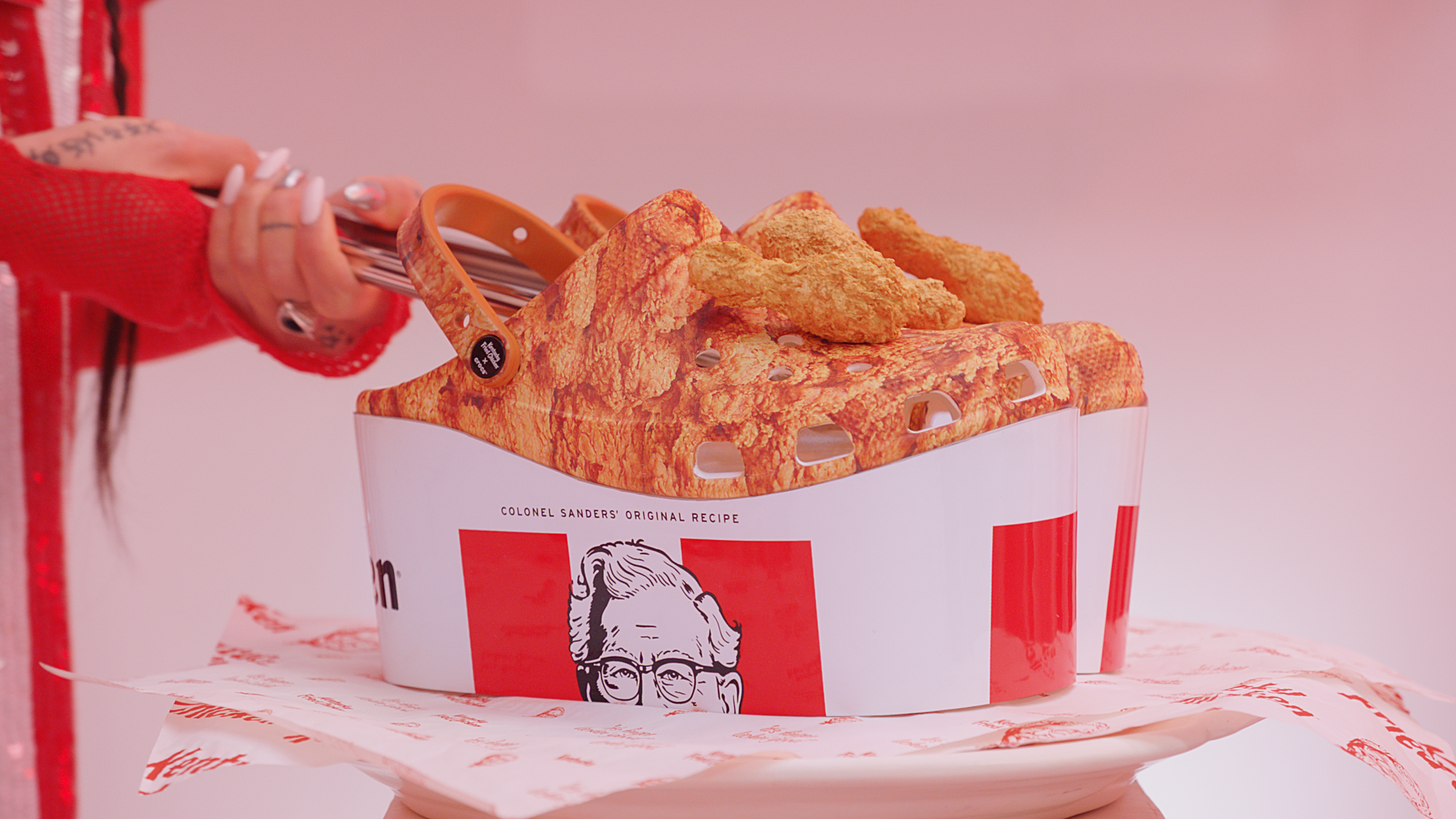 A container of KFC fried chicken with Colonel Sanders logo, held by a person's hand with long painted nails, against a pink background.