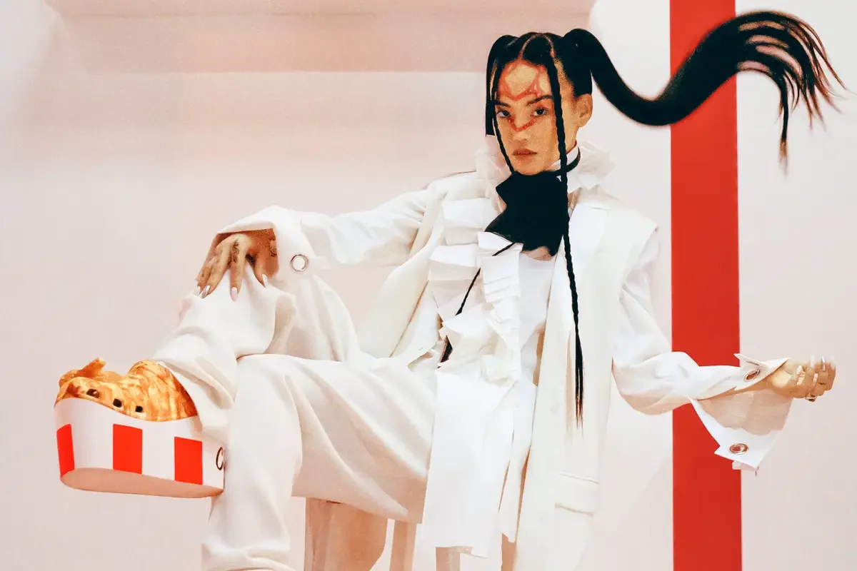 Person in white clothing with long black braided hair holding a box of french fries, posing in front of a red and white background.