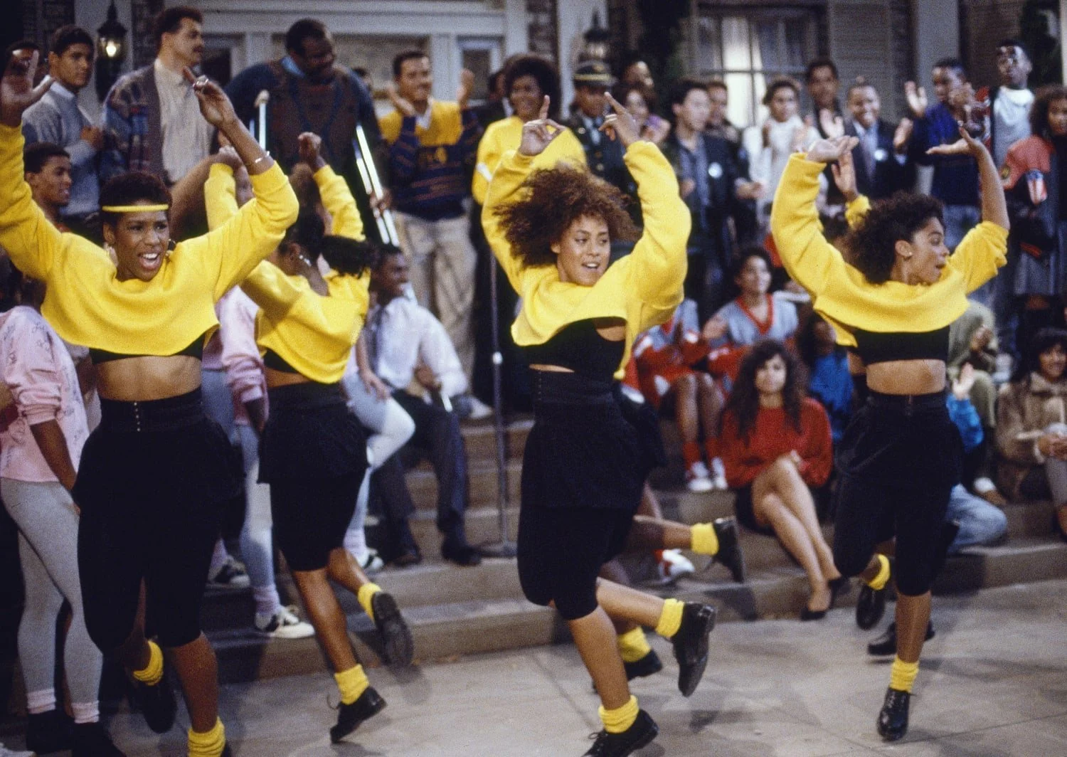 Three women dancing in coordinated yellow and black outfits with an audience watching outdoors at night.