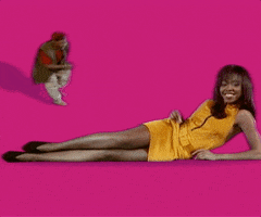 A woman in a yellow dress lying on her side on a pink background, smiling, with a small monkey sitting in a relaxed position nearby.