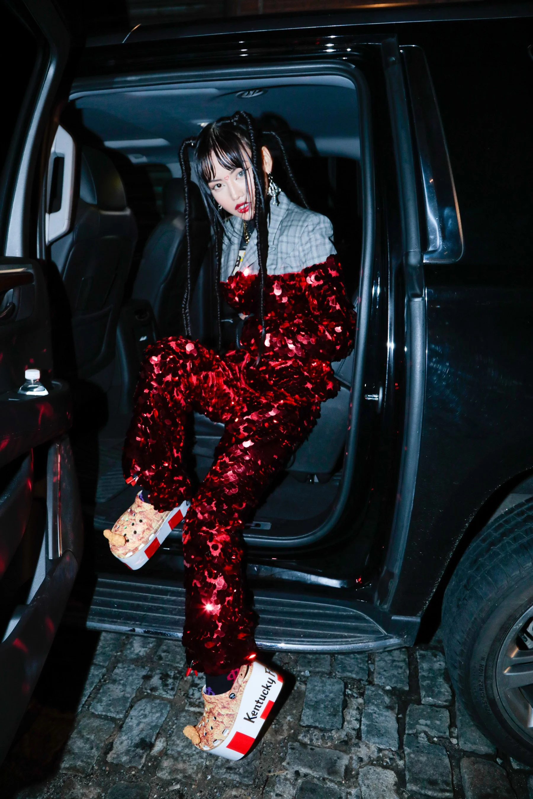 A woman with black braided hair and makeup, sitting in the back of a vehicle, wearing a sparkly red and gray outfit, holding a pizza box, with the mark 'Kentucky' visible on the box.