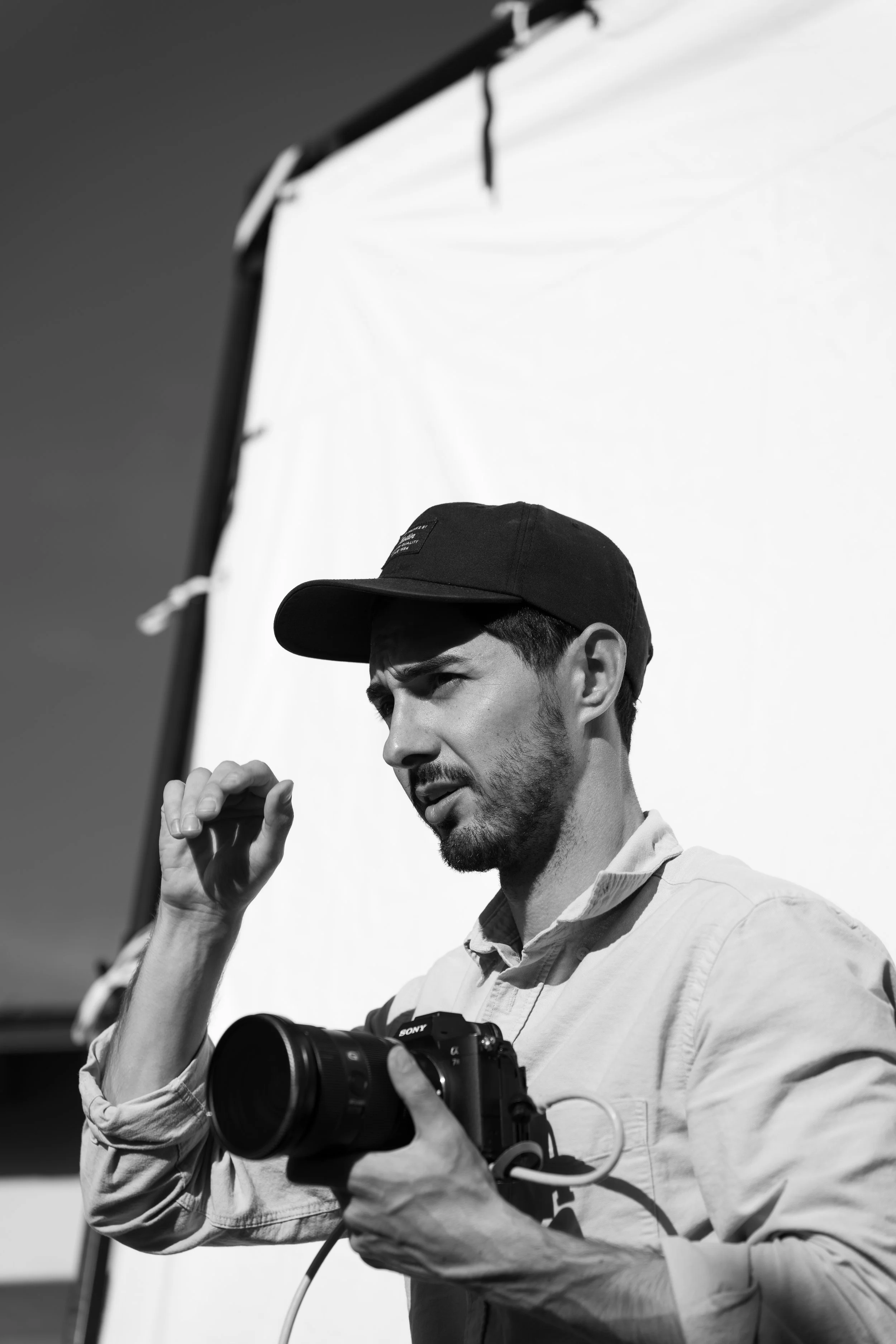 A man is holding a Sony camera and adjusting his cap while standing in front of a white backdrop during a photo shoot.