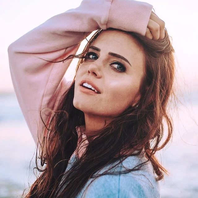 Musician Tiffany Alvord with long, wavy brown hair and blue eyes posing outdoors at the beach during sunset, wearing a pink hoodie.