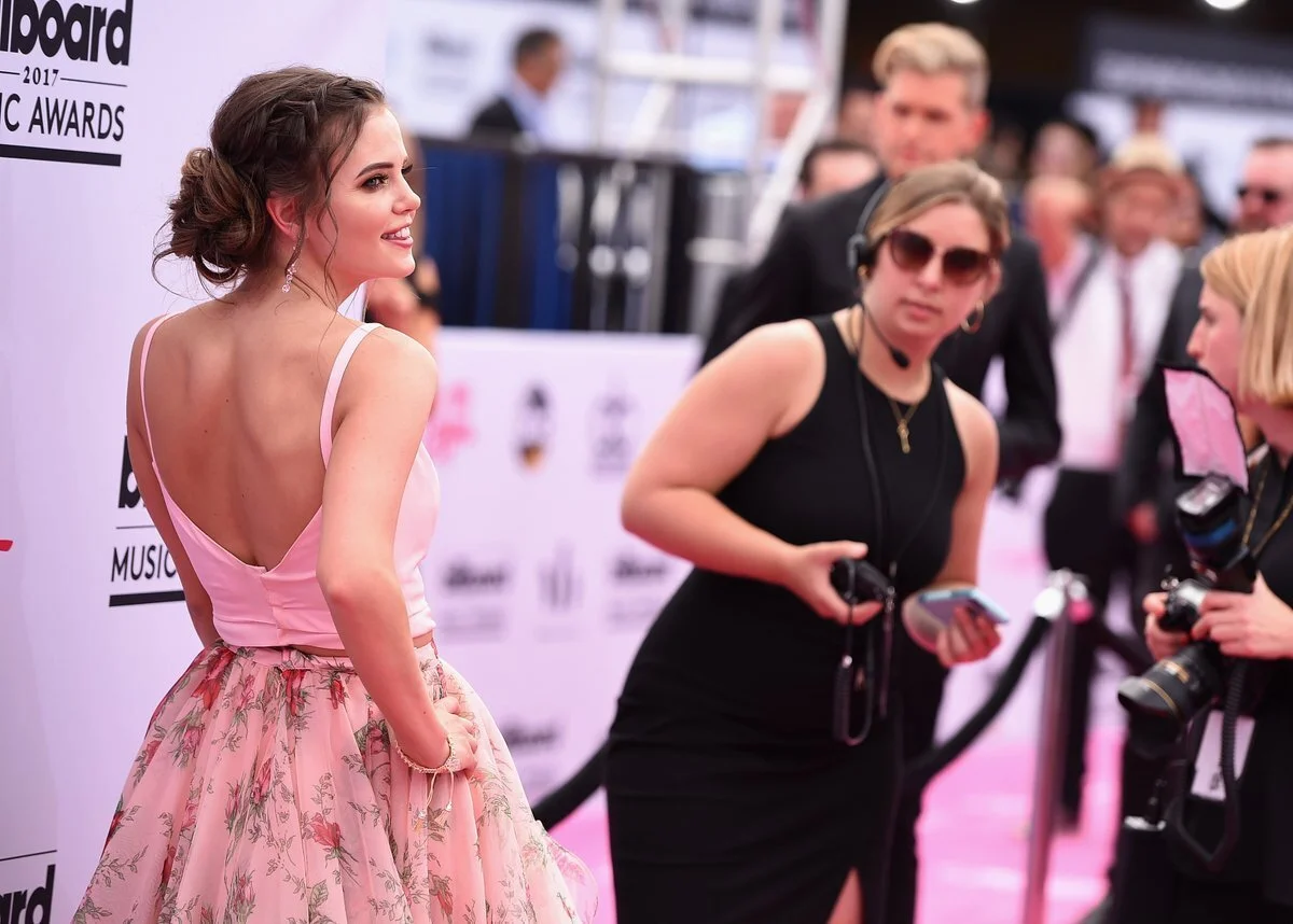 Tiffany Alvord in a pink satin top and floral skirt posing on a pink carpet at the Billboard Music Awards, being photographed and interviewed by media personnel.