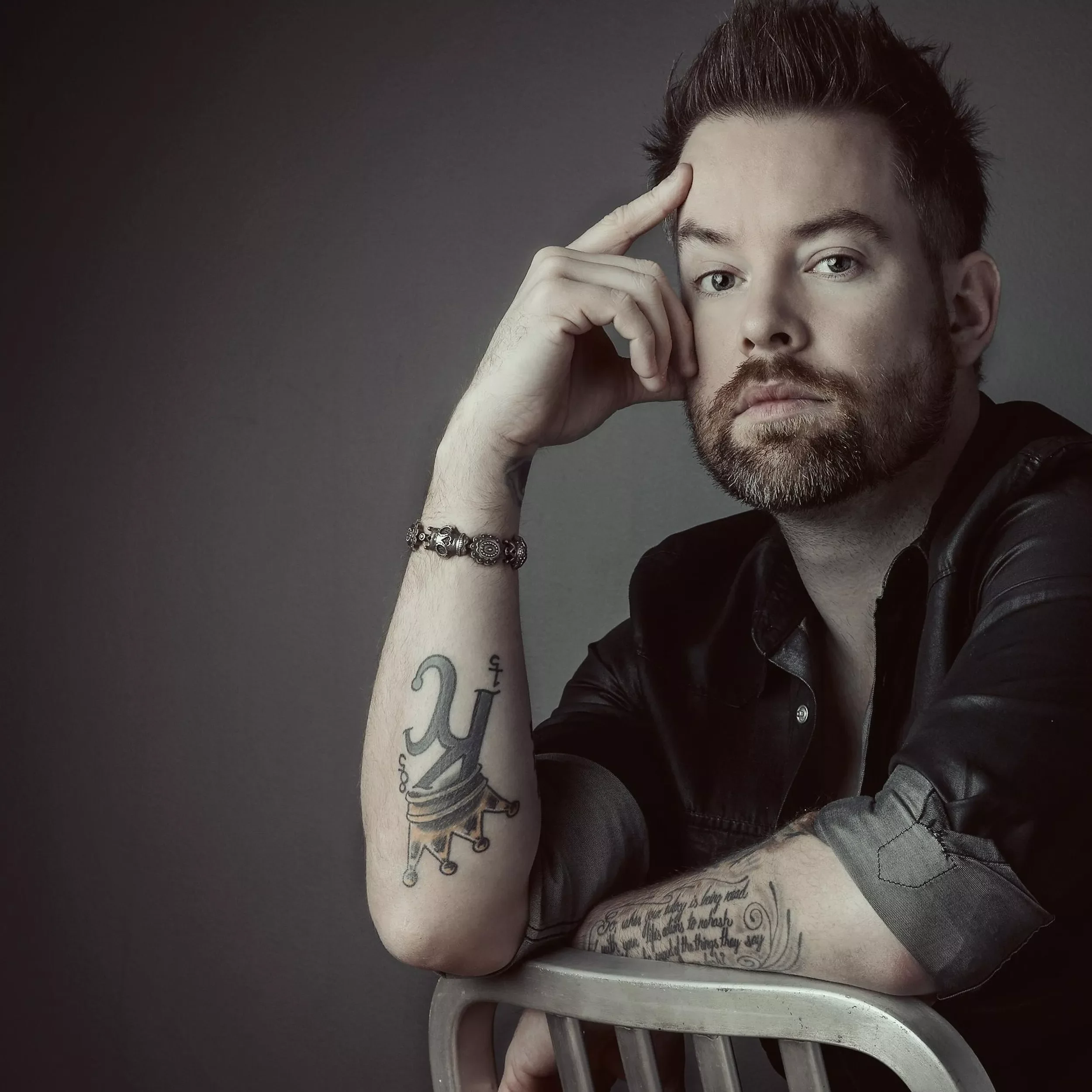Musician David Cook with a beard and short brown hair, holding a metallic mug, is leaning on a metal chair with his left arm. His right hand is resting on his forehead, and he is looking at the camera.
