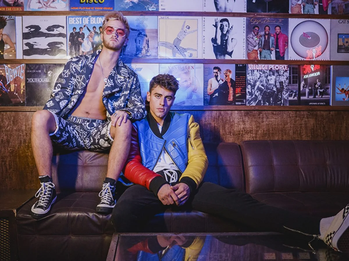 Music duo Jack & Jack posing in a retro music lounge with vinyl records on the wall behind them. One sitting on the sofa wearing a colorful jacket and black pants, the other sitting on a high stool wearing sunglasses,