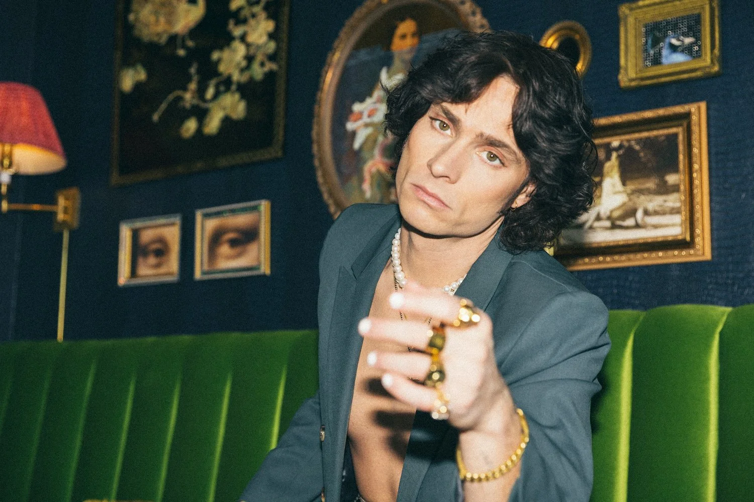 Musician Spencer Sutherland with dark, wavy hair and light skin sitting on a green velvet couch, wearing a gray blazer, pearl jewelry, and rings, pointing toward the camera with a serious expression.