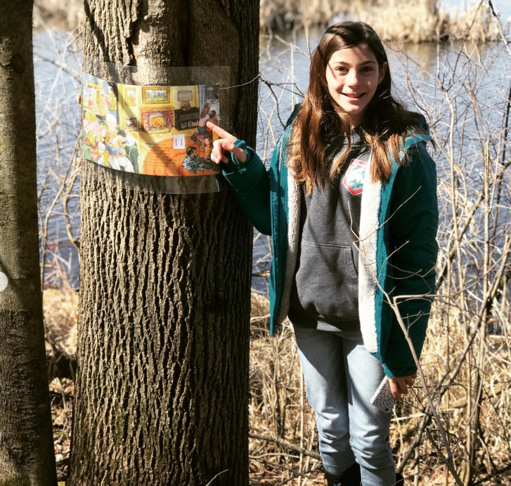 A girl pointing to an inner page of "Under in the Mud" on a tree during a StoryWalk