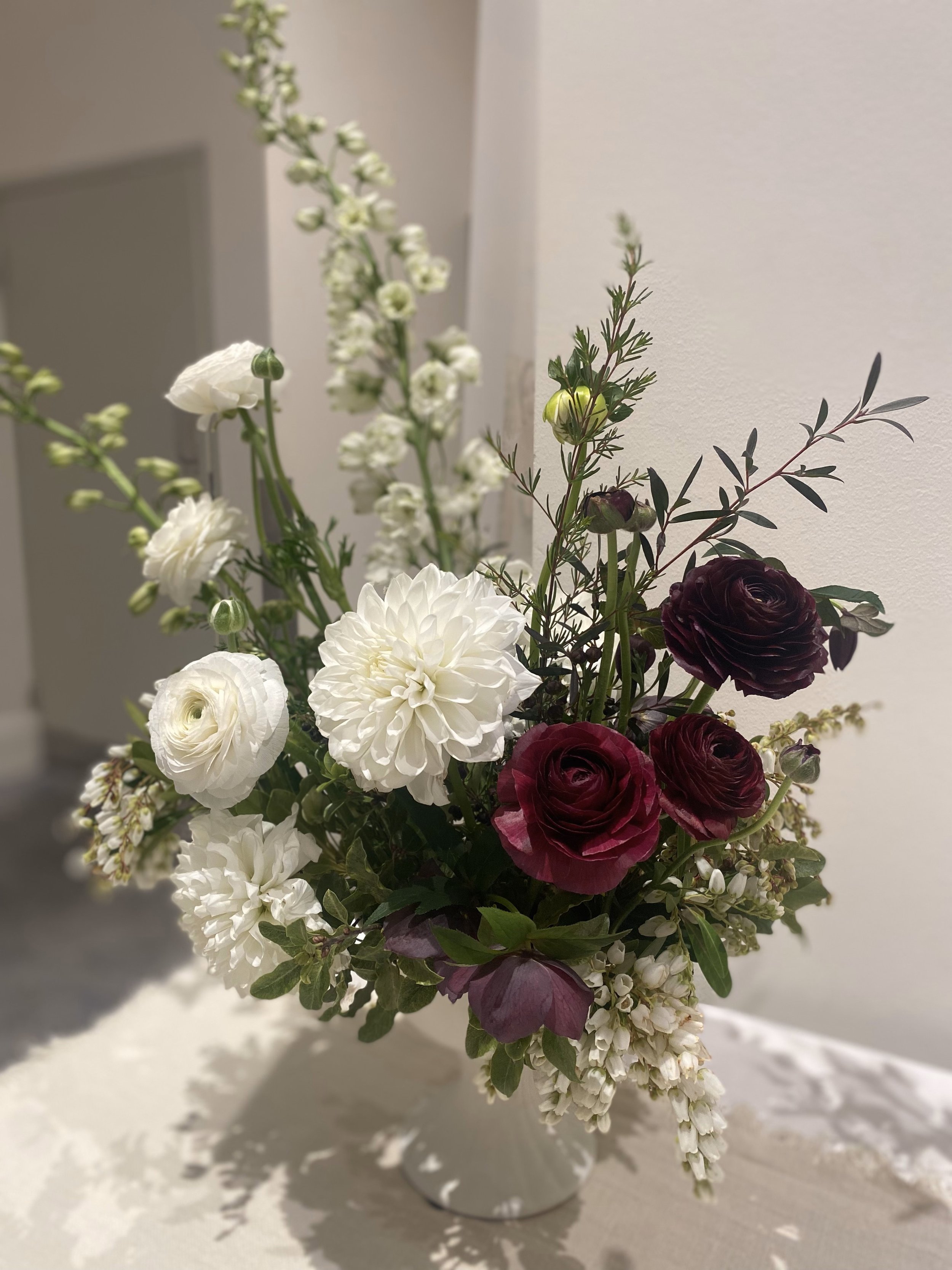 A sophisticated floral compote arrangement filled with tall spires of white delphinium, rich burgundy ranunculus and helleborus, as well as white dahlias, ranunculus and flowing dainty andromeda flowers. A stunning statement floral design by Hart and