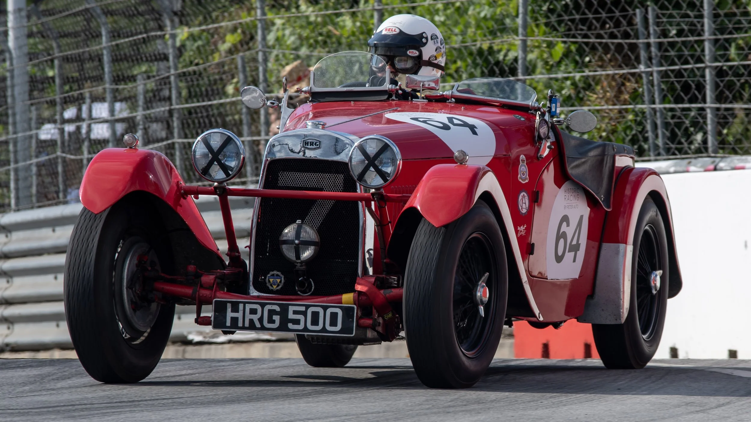 Red vintage race car with number 64, driven by a helmeted driver on a racetrack, surrounded by safety fencing.