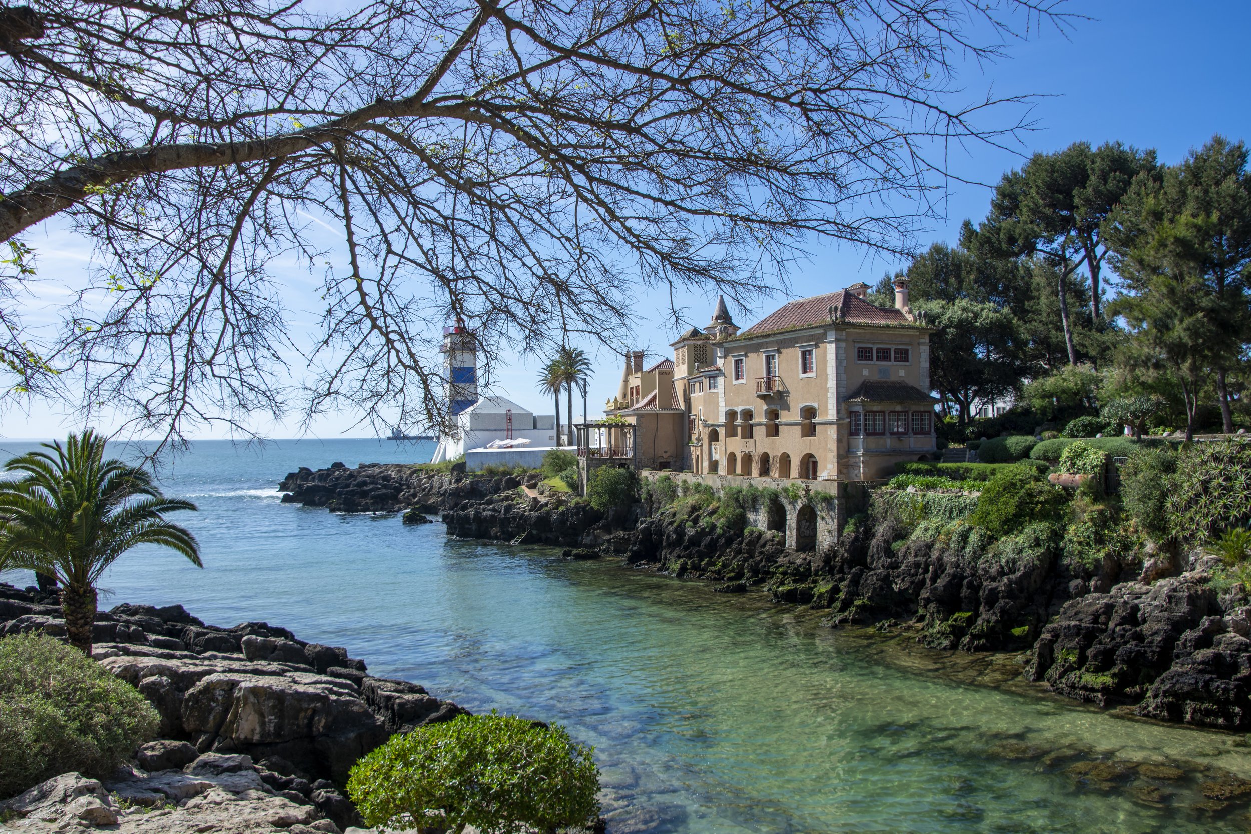 Scenic coastal view with a historic building, lighthouse, and lush trees. The foreground features rocky terrain and a clear, calm ocean under a bright blue sky.