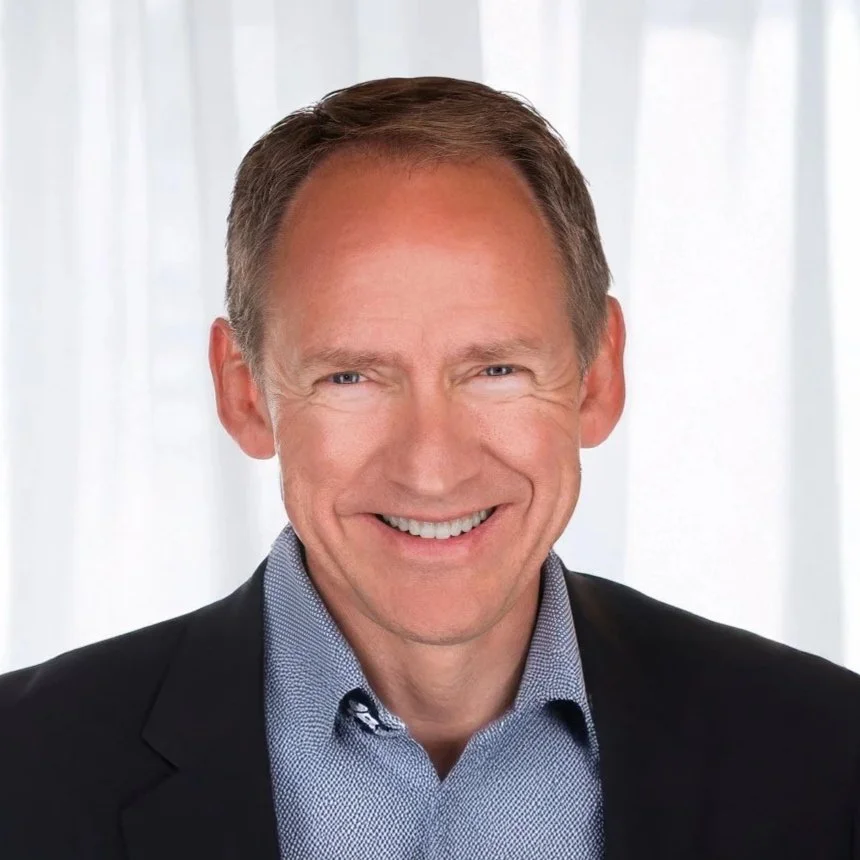 Portrait of a smiling middle-aged man with short brown hair, wearing a dark suit and light blue dress shirt, standing in front of a light-colored curtain background.