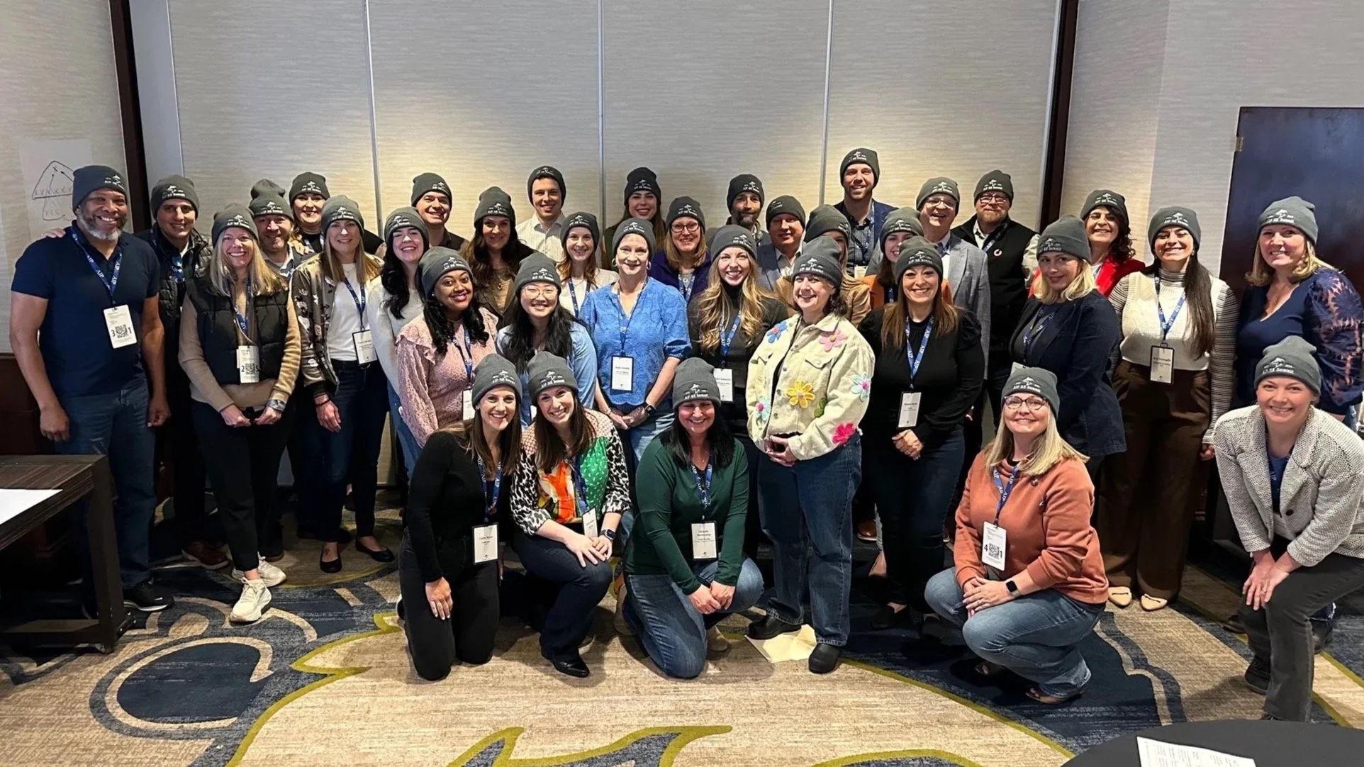 A group of diverse people posing together in a conference room, all wearing matching gray beanie hats with a logo, smiling for the camera.