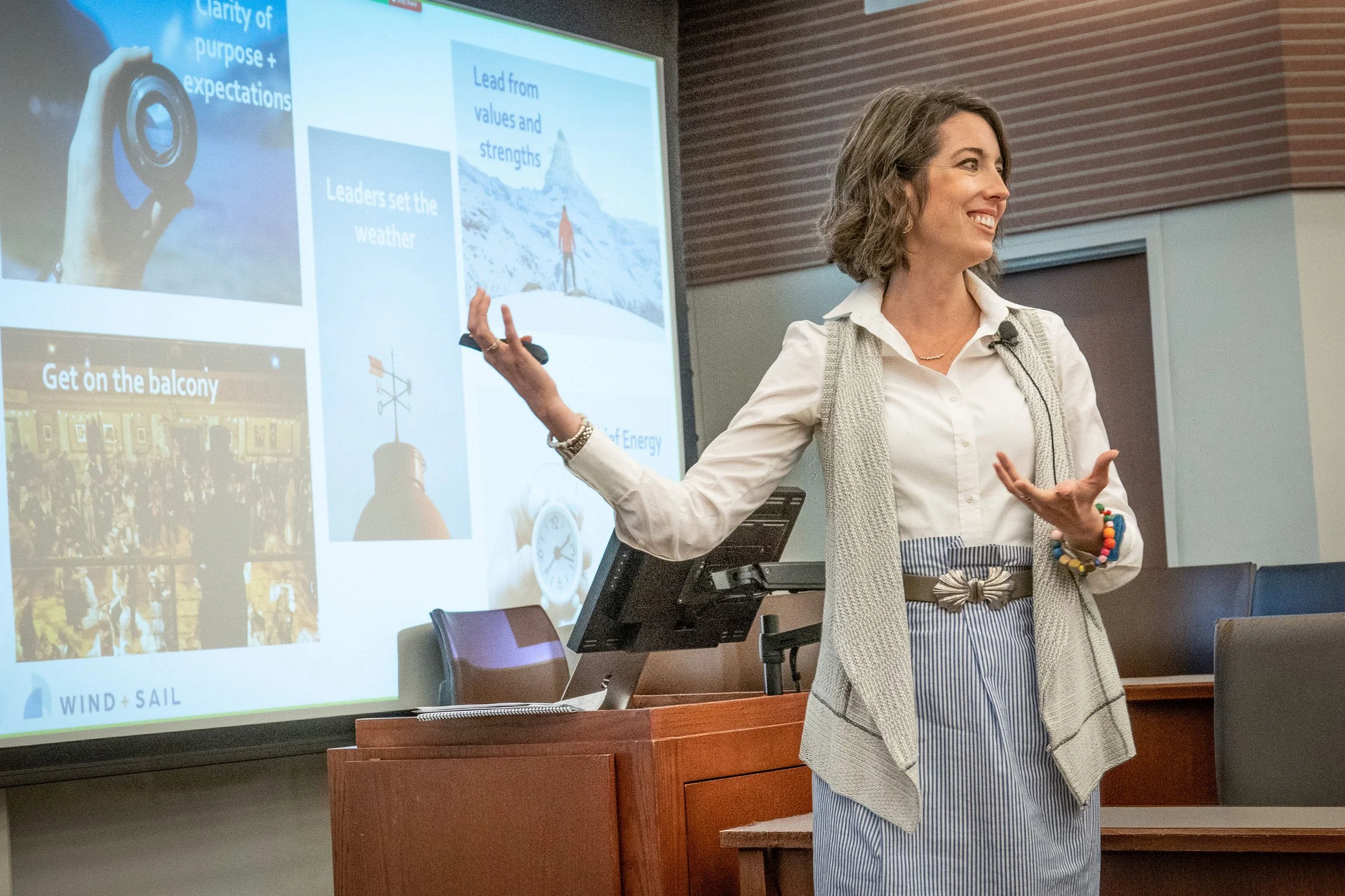 A woman giving a presentation in a conference room with a slide projected behind her. She is smiling, wearing a white shirt, beige vest, and striped pants. The slide contains text and images related to leadership, purpose, and expectations.